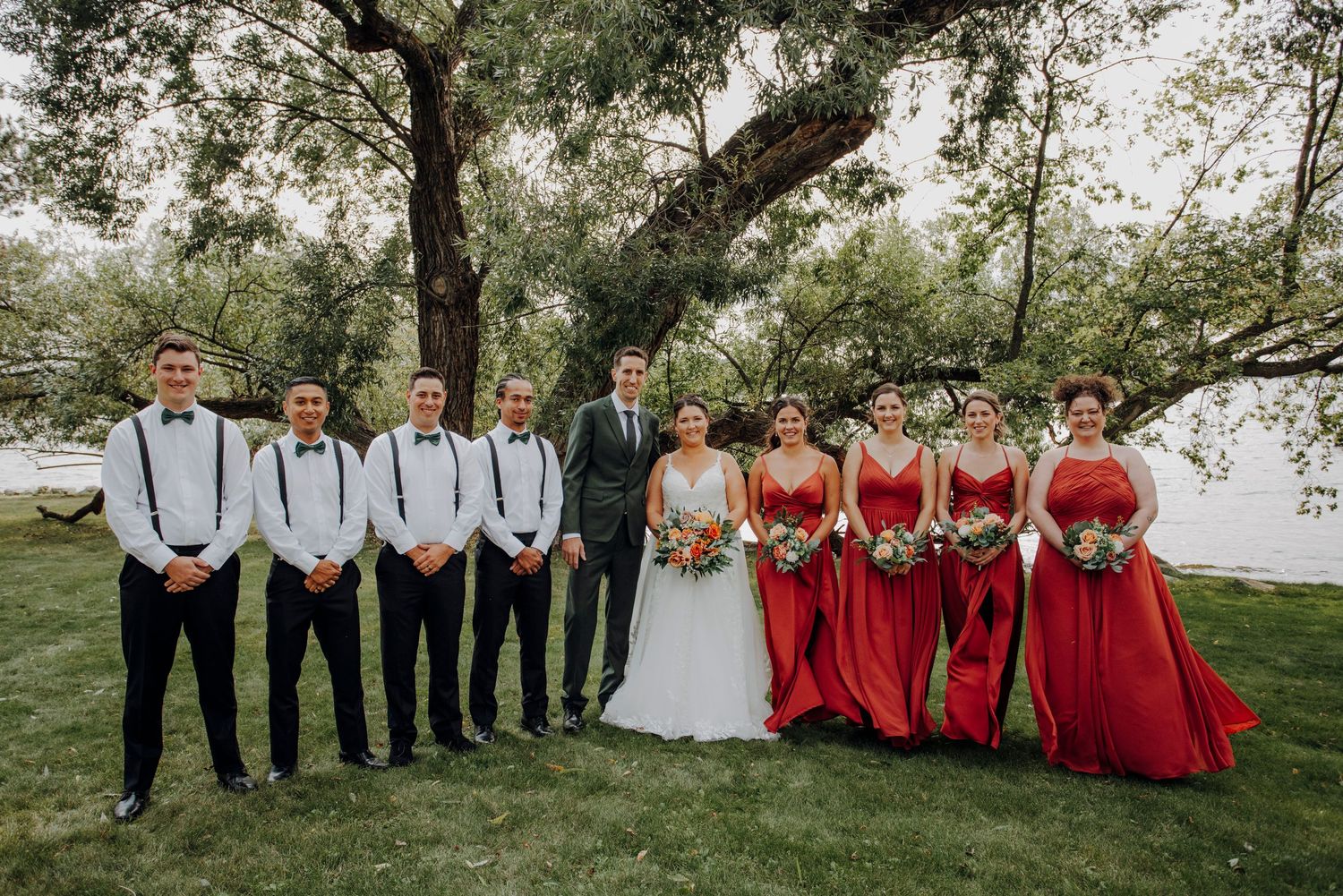 Wedding party in red bridesmaid dresses and groomsmen in dark suits pose together outdoors.