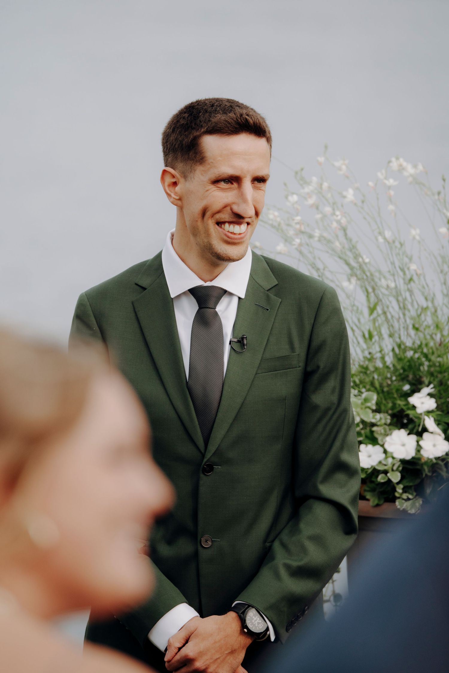 Groom in dark suit smiles warmly against grey background with white floral decor.