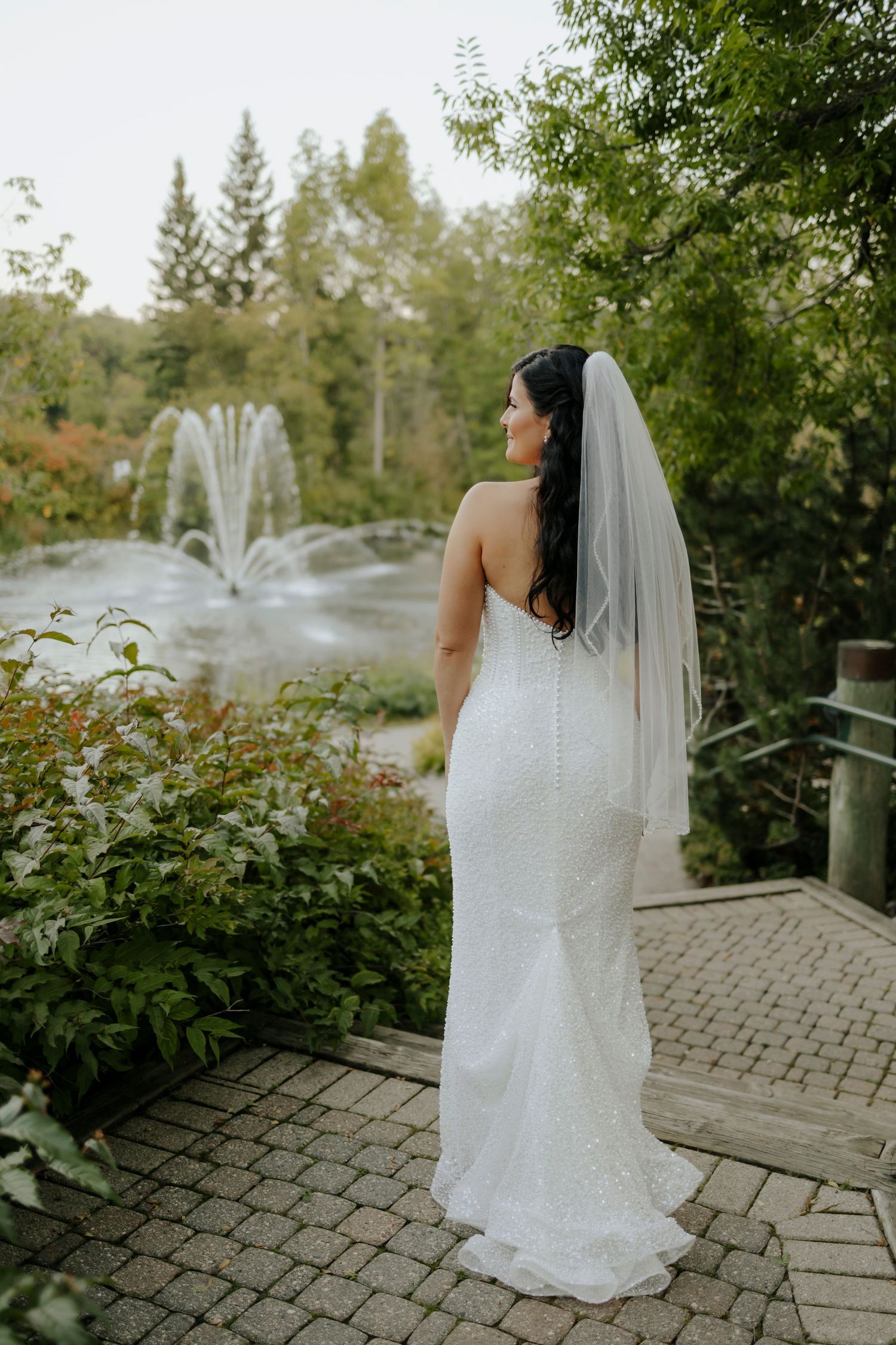 A bride in a fitted white dress stands near a fountain, photographed from behind.