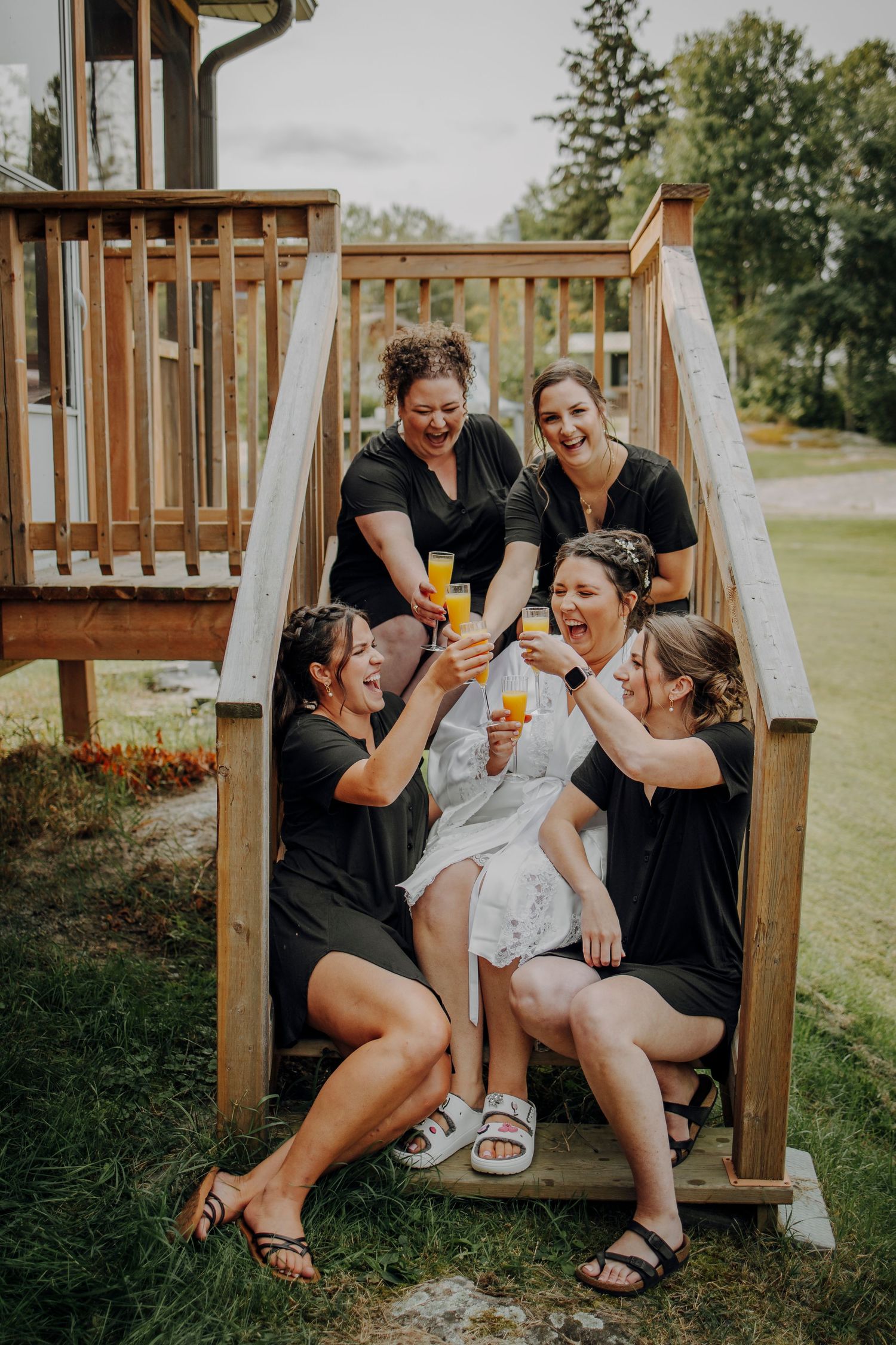 Wedding party in black attire sharing drinks and laughter on outdoor wooden stairs.