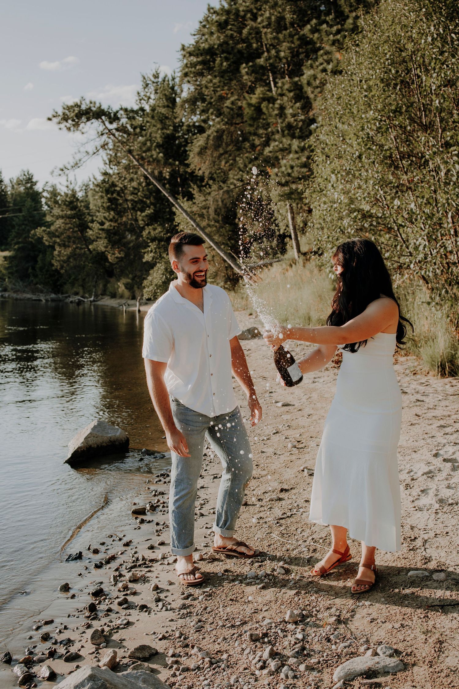 Playful couple popping champagne on a rocky lakeshore during their engagement celebration.