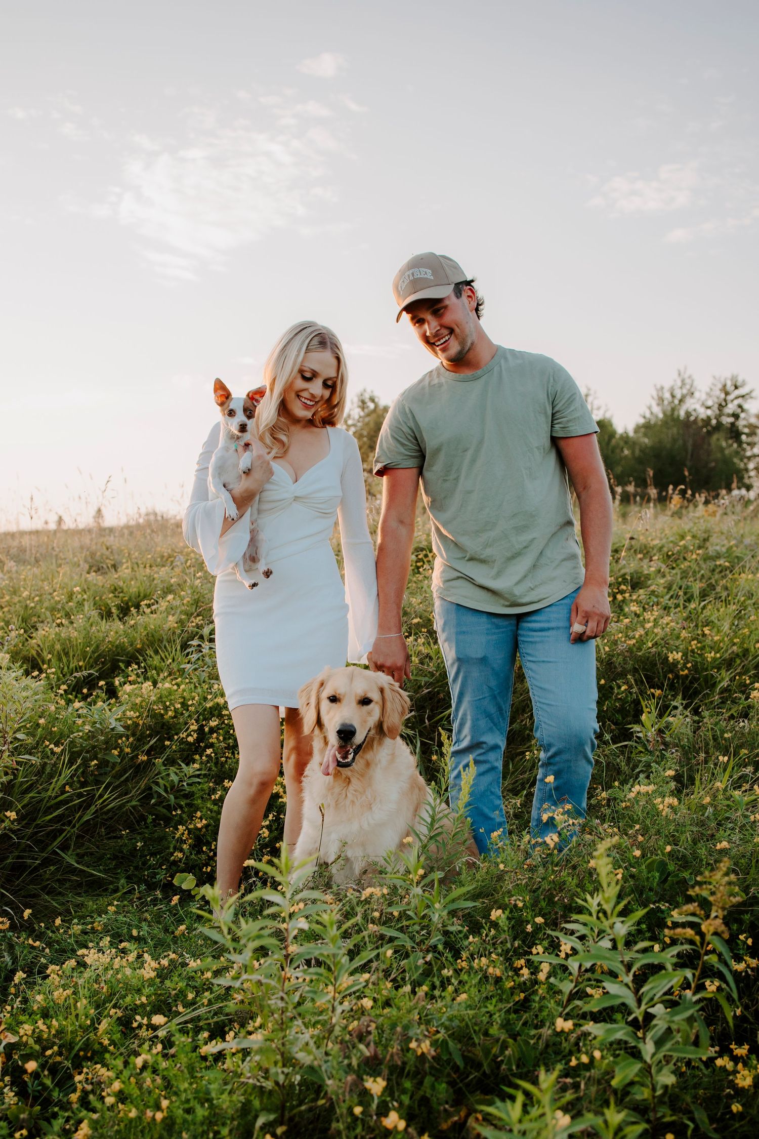 A couple walks their golden retriever and small dog through a sunny meadow.