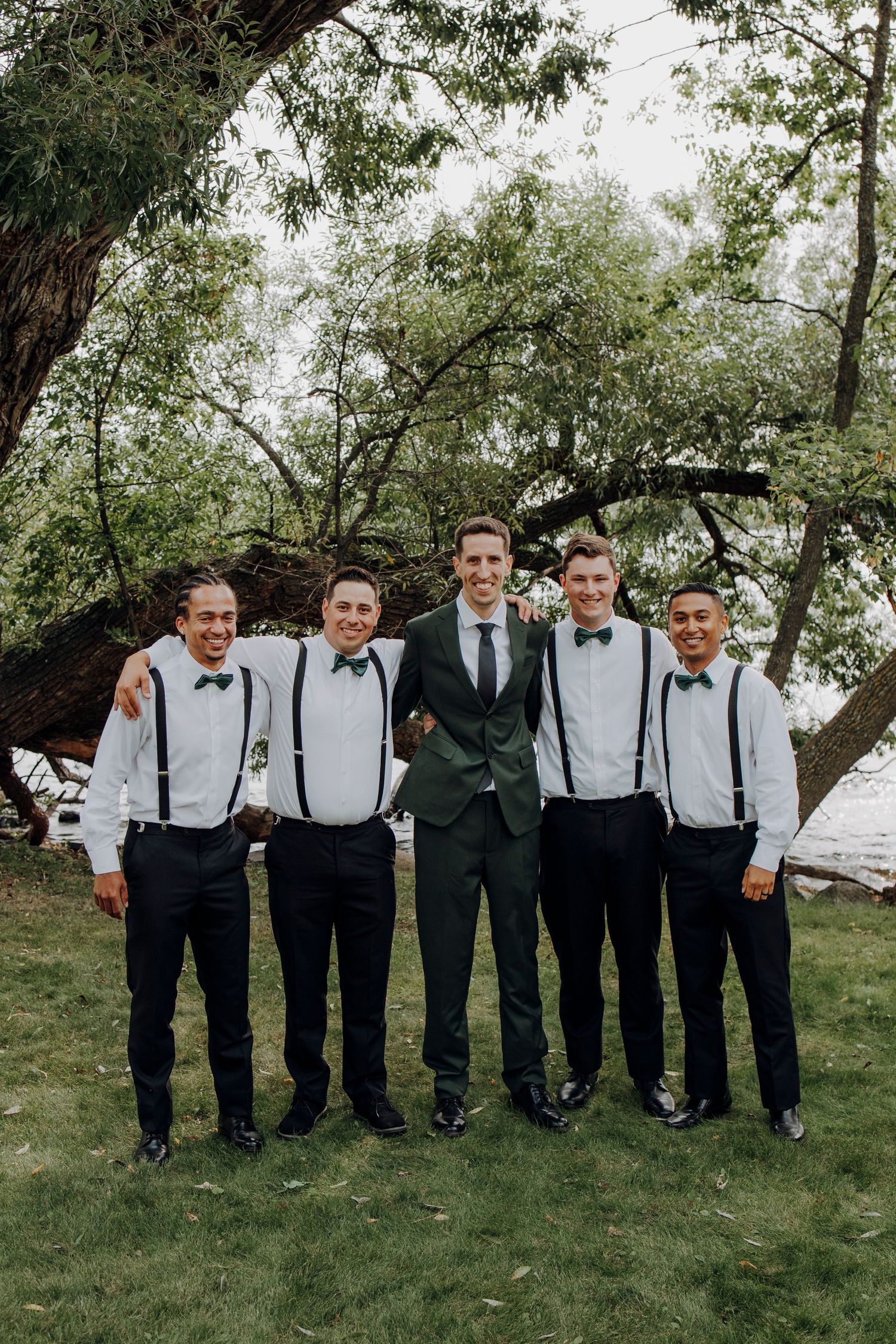 Groomsmen in suspenders and bowties pose together on a green lawn.