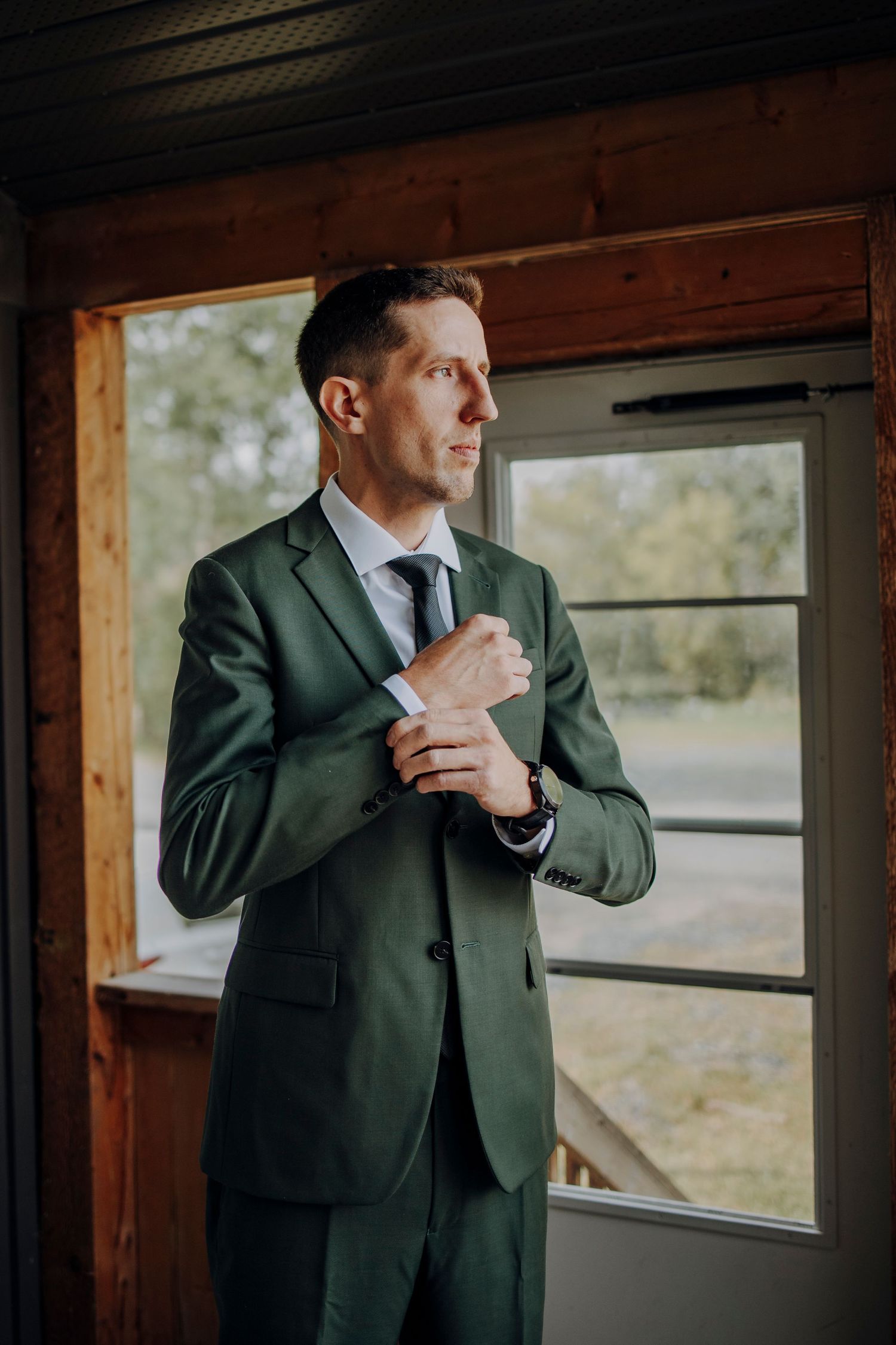 A groom in a dark green suit jacket adjusts his tie while standing in a rustic wooden building.