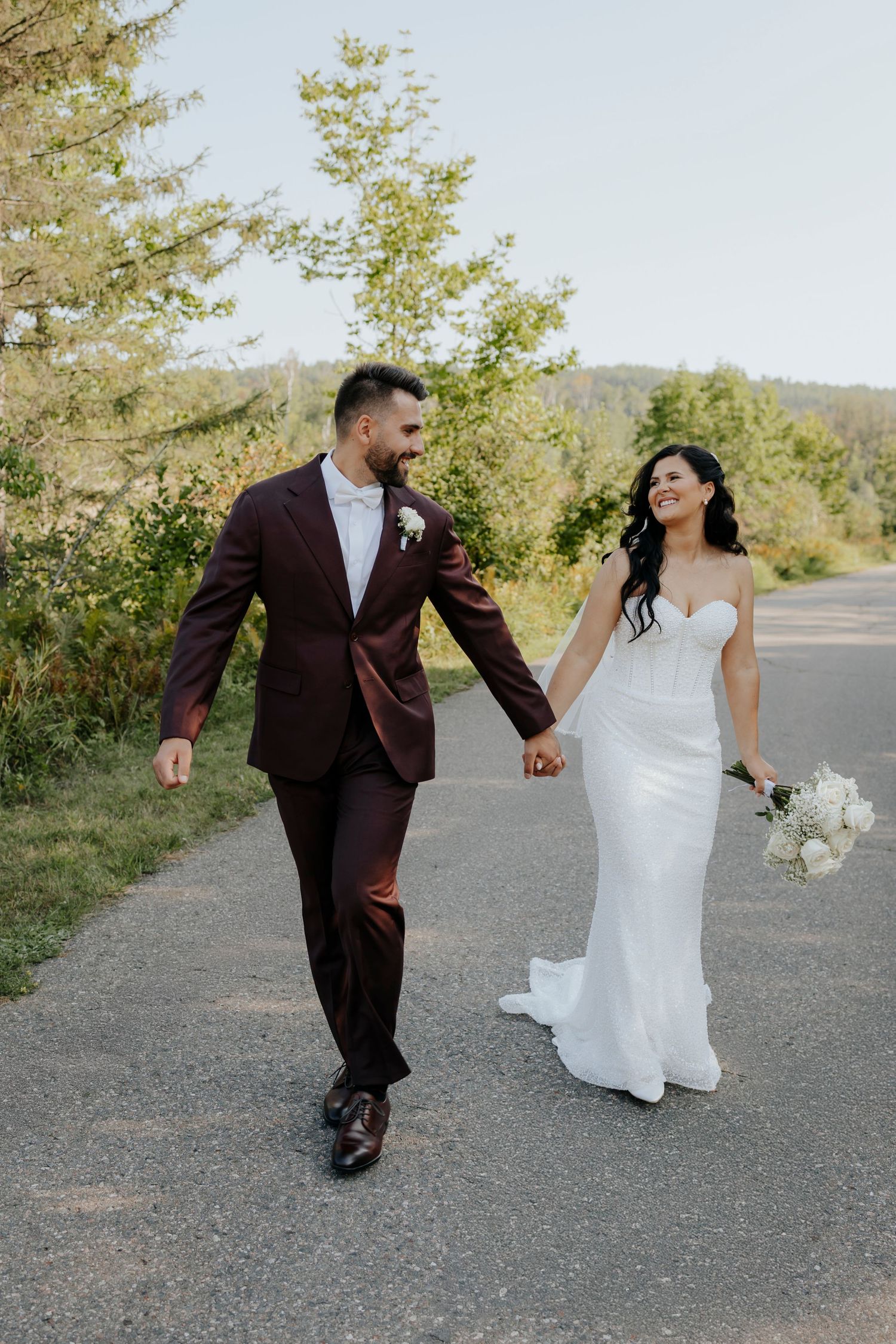 A couple in wedding attire walks playfully along a gravel path with trees and mountains in the background.