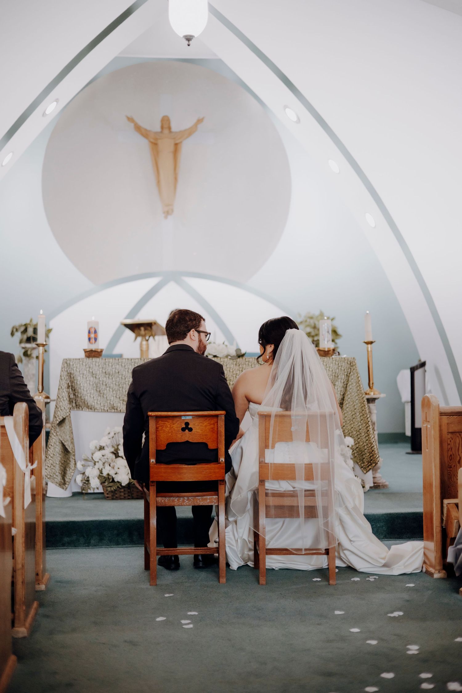 A wedding ceremony in a small white chapel with wooden pews and a simple crucifix on the wall.