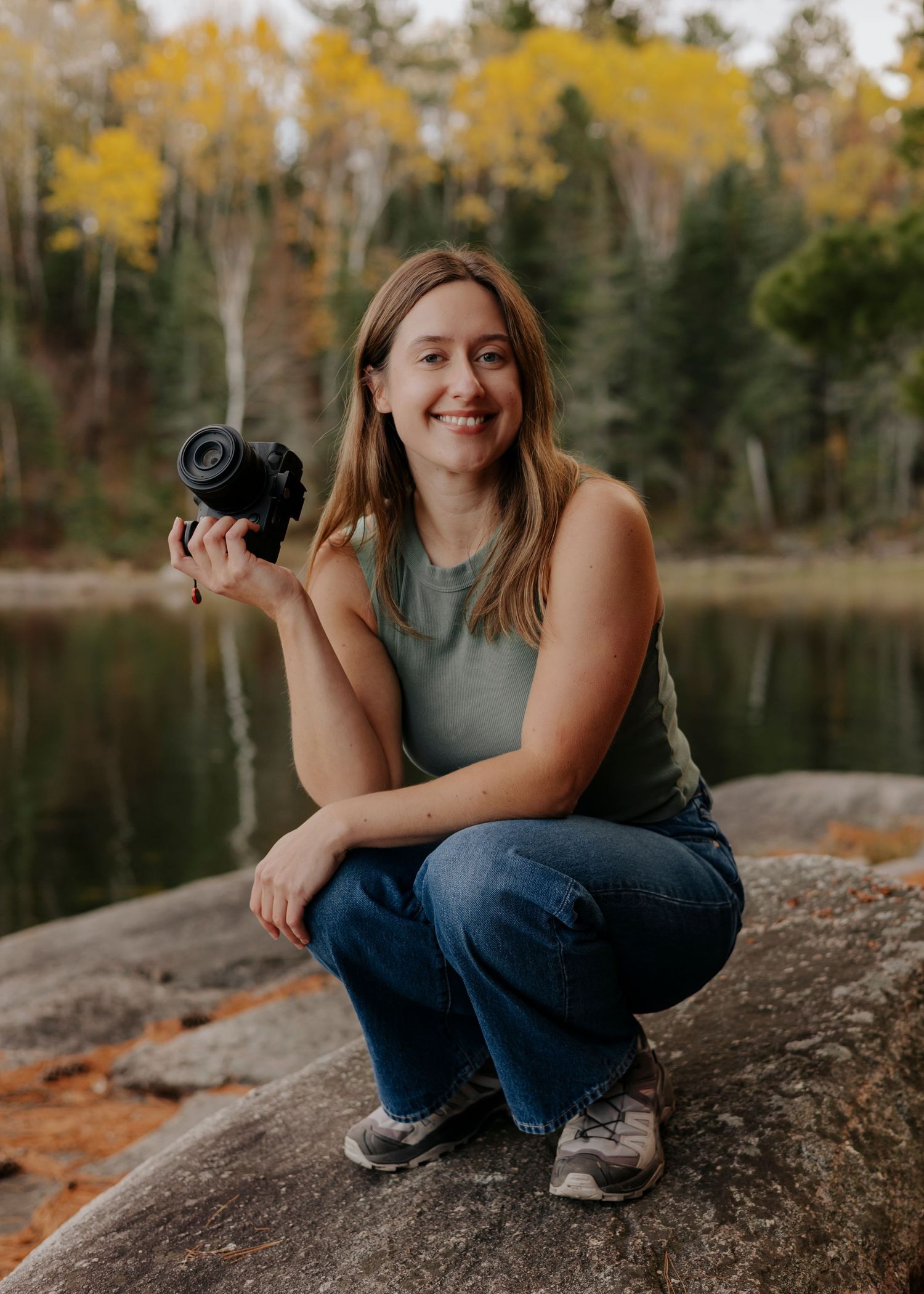 Woman photographer smiling, crouching on rocks holding camera, surrounded by autumn trees and lake backdrop.