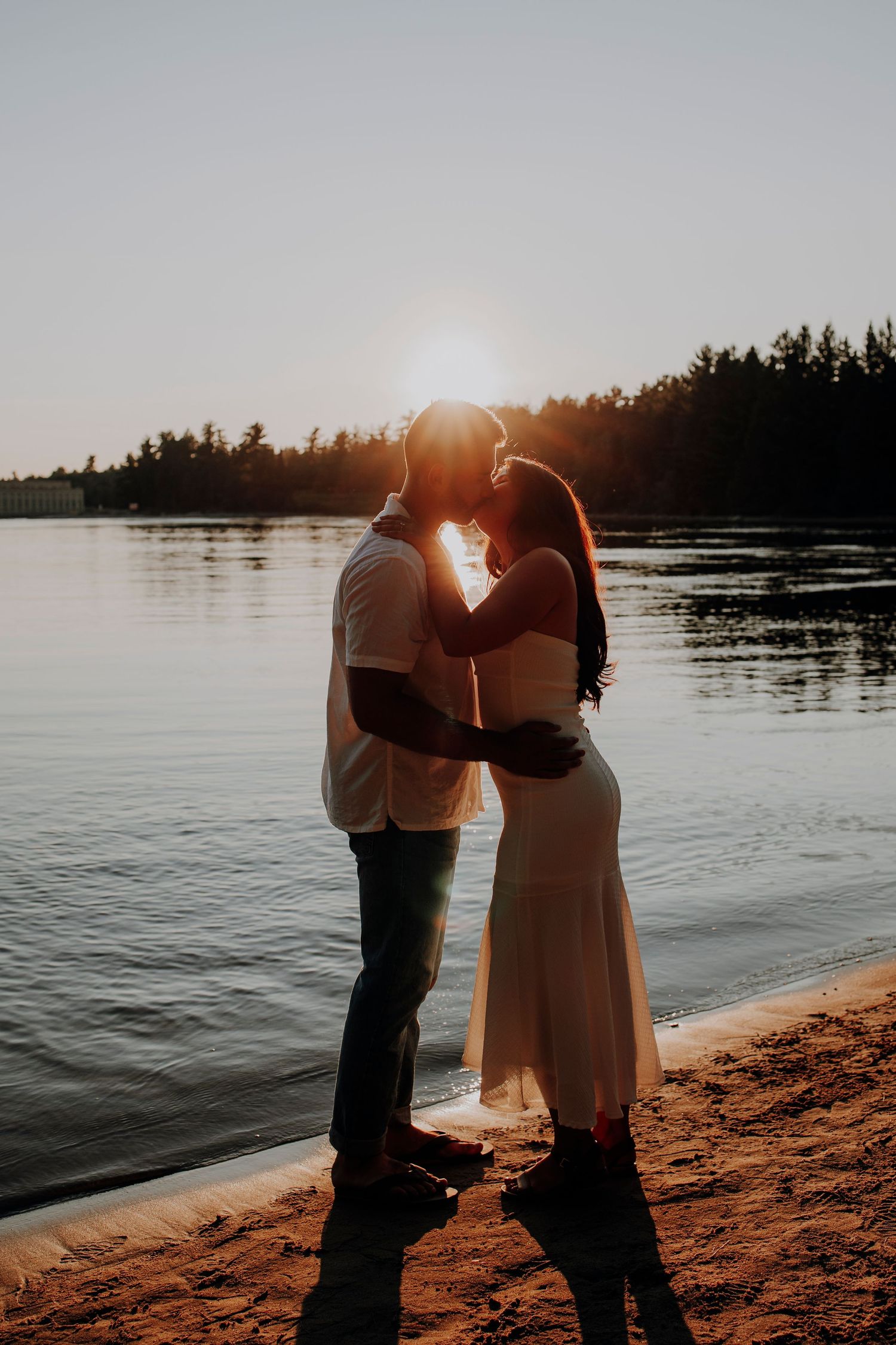 Romantic sunset silhouette of a couple embracing on a beach with a lake and forest backdrop.