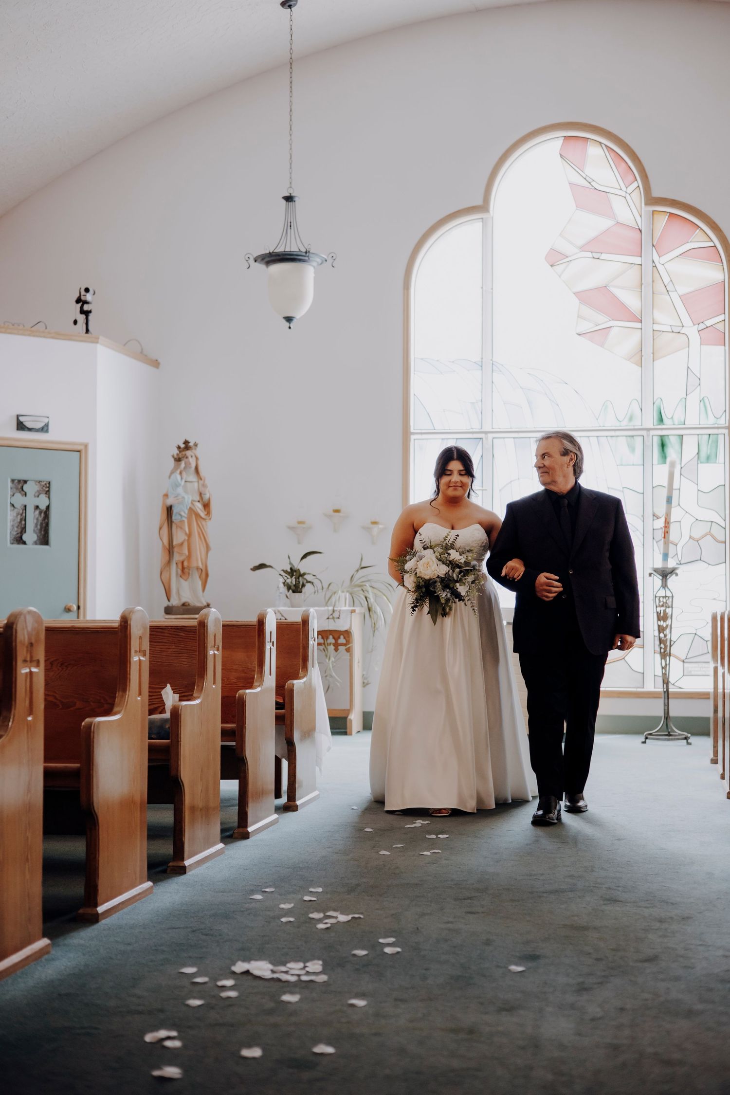 A church aisle with wooden pews and arched windows features scattered rose petals on the floor.