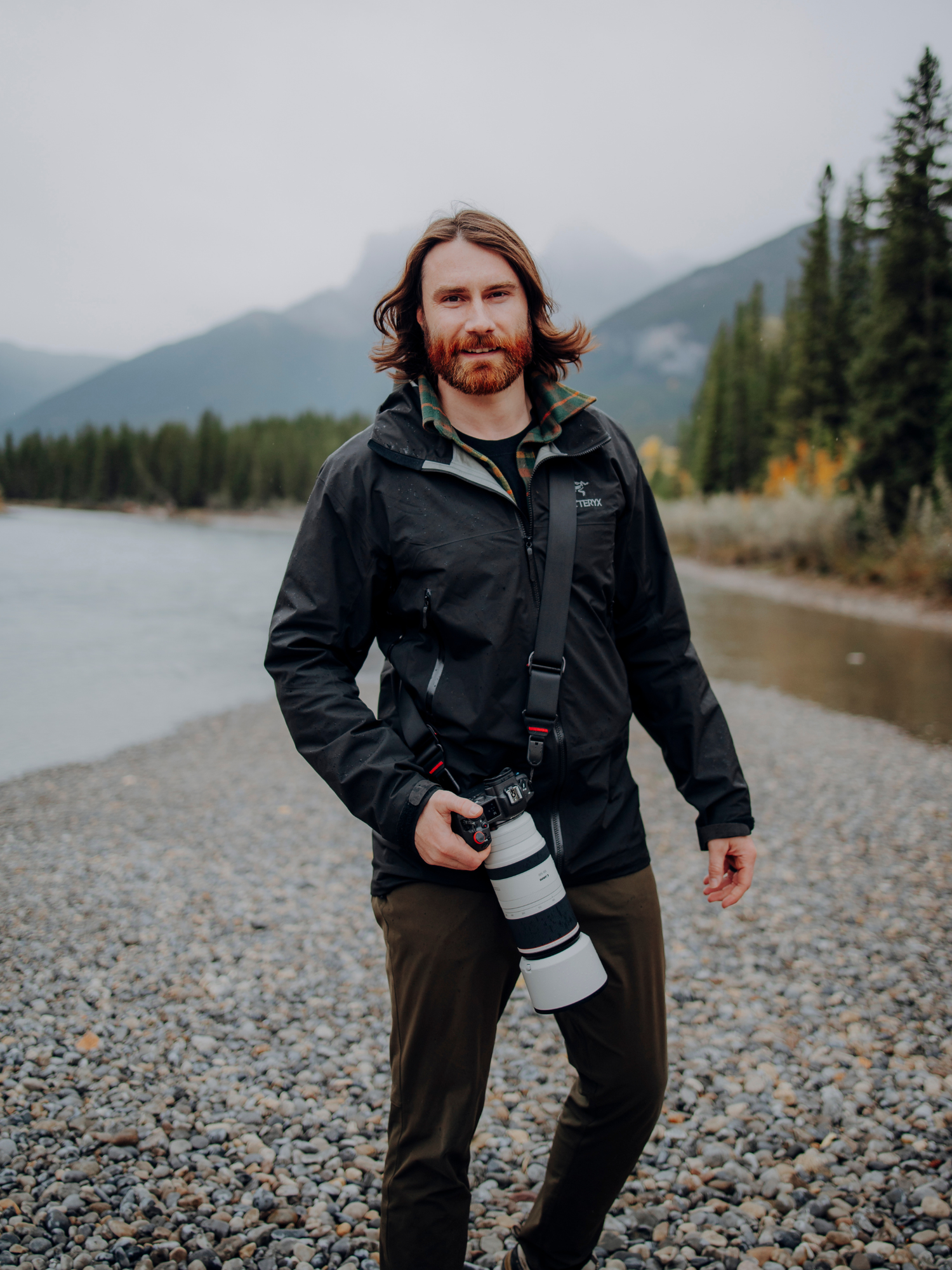 Photographer with telephoto lens standing on rocky shoreline against mountain landscape.