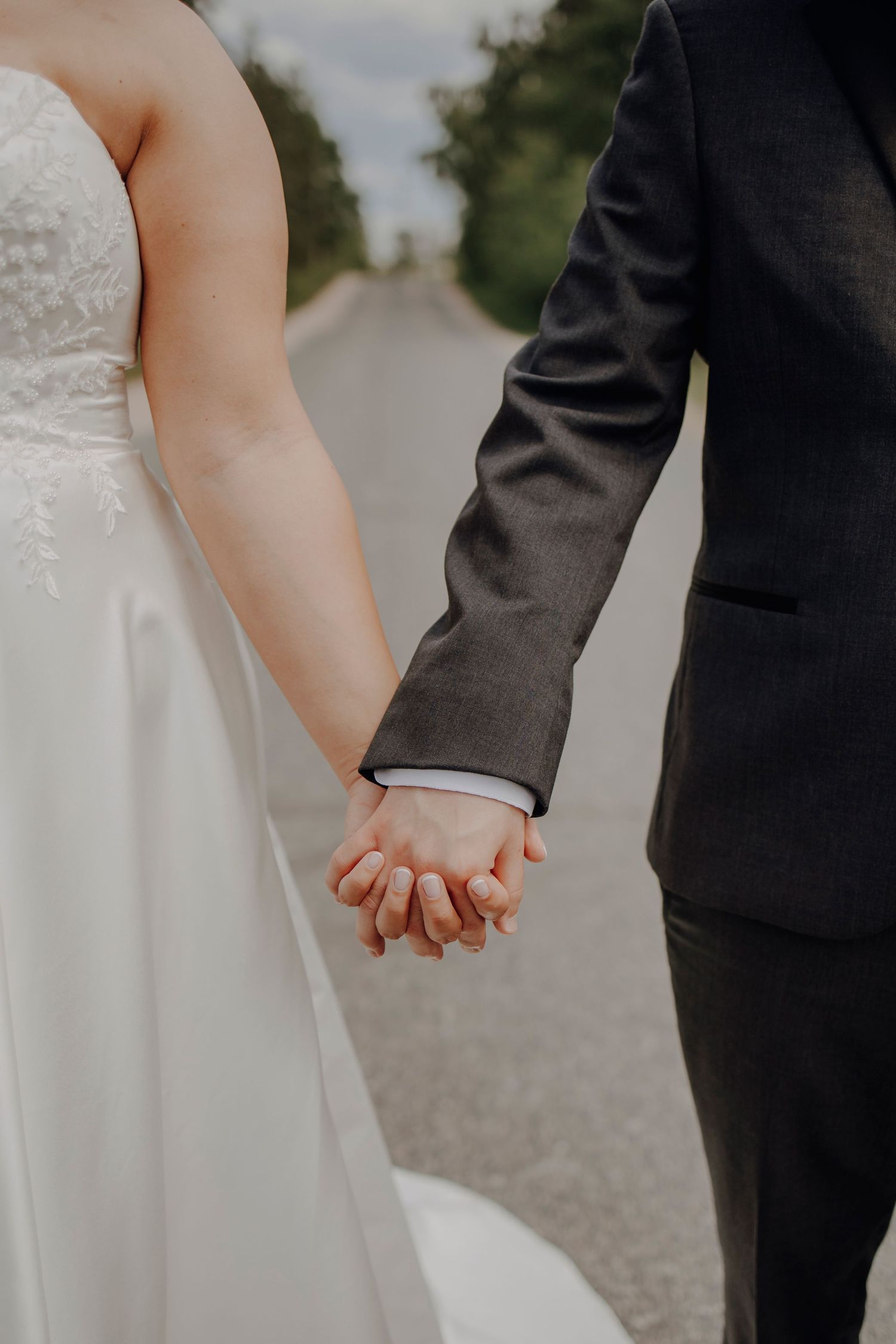 A close-up of newlyweds holding hands on a tree-lined path, with bride in white dress and groom in dark suit.