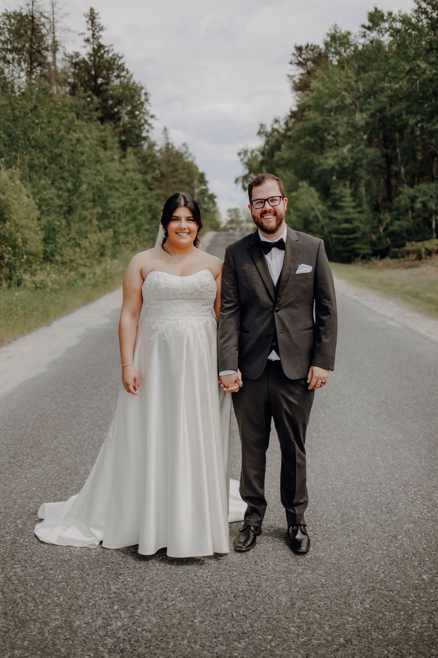 A wedding couple poses playfully on a country road surrounded by green trees.