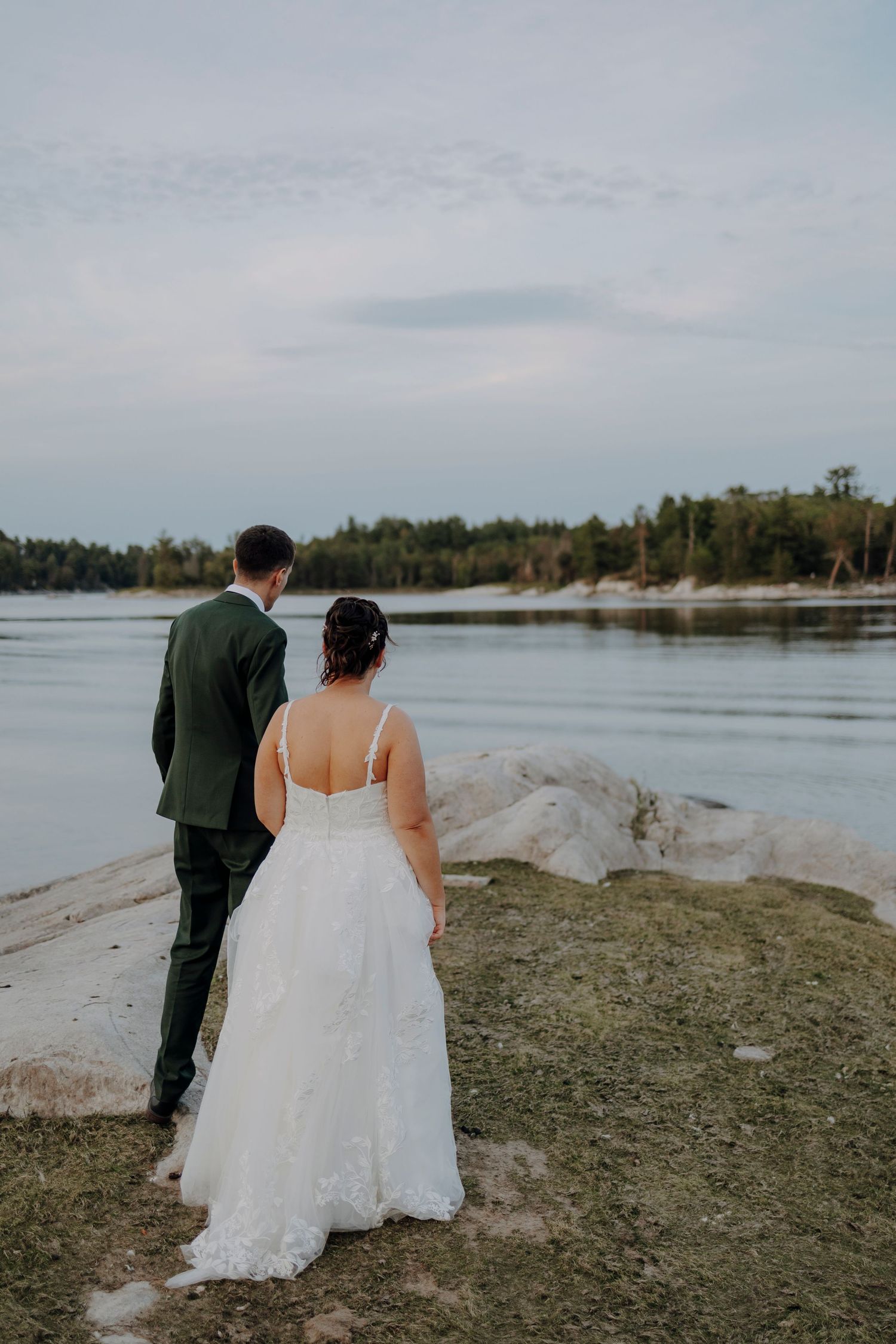 Newlywed couple walks along a scenic waterfront with trees and cloudy skies in the background.