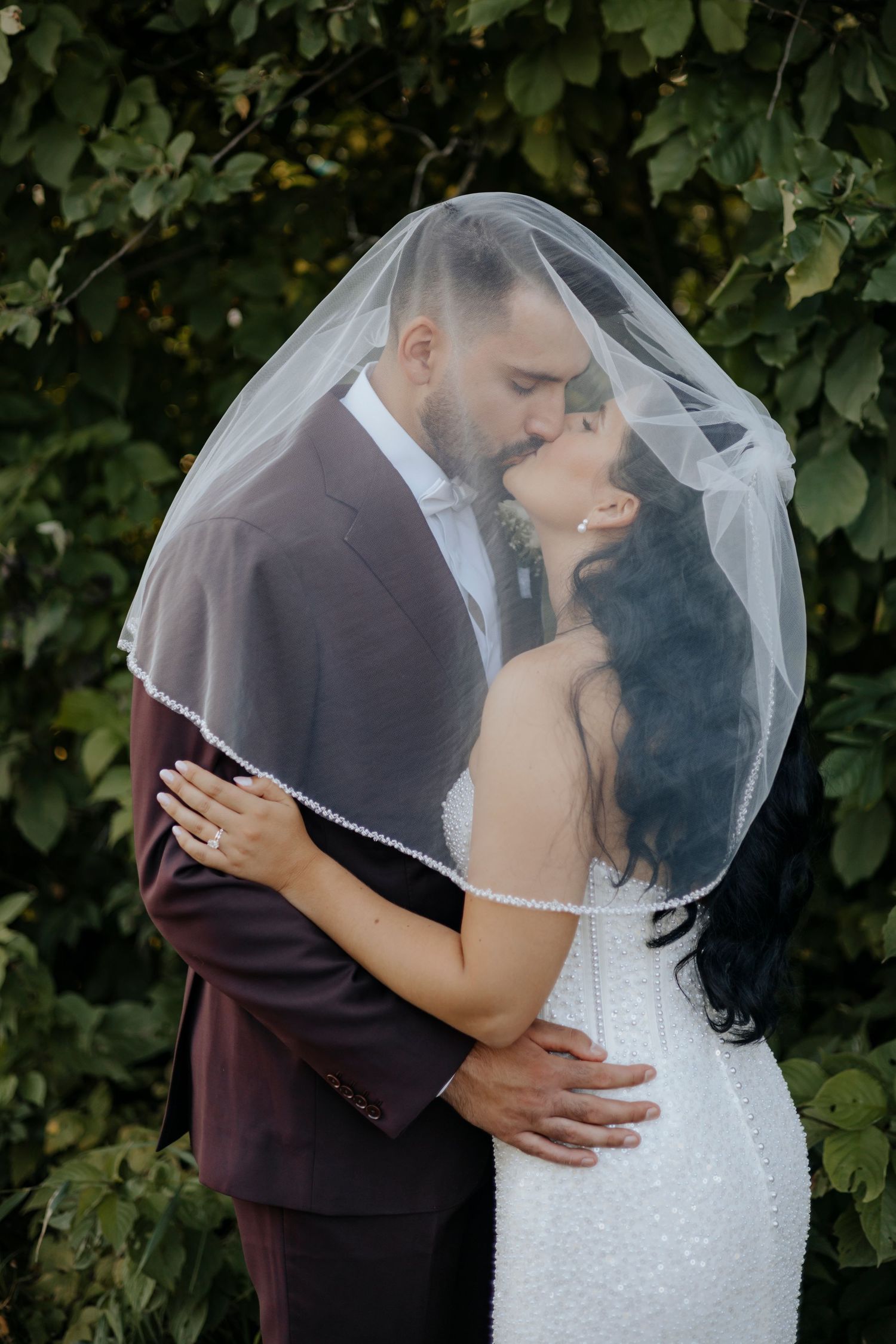 A romantic wedding couple shares intimate kisses under a delicate veil against a leafy green backdrop.
