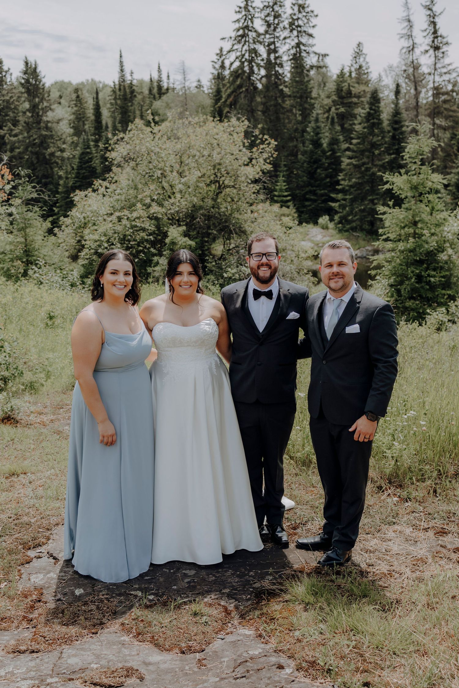 Wedding party in formal attire poses together on a mountain path surrounded by evergreen trees and natural scenery.