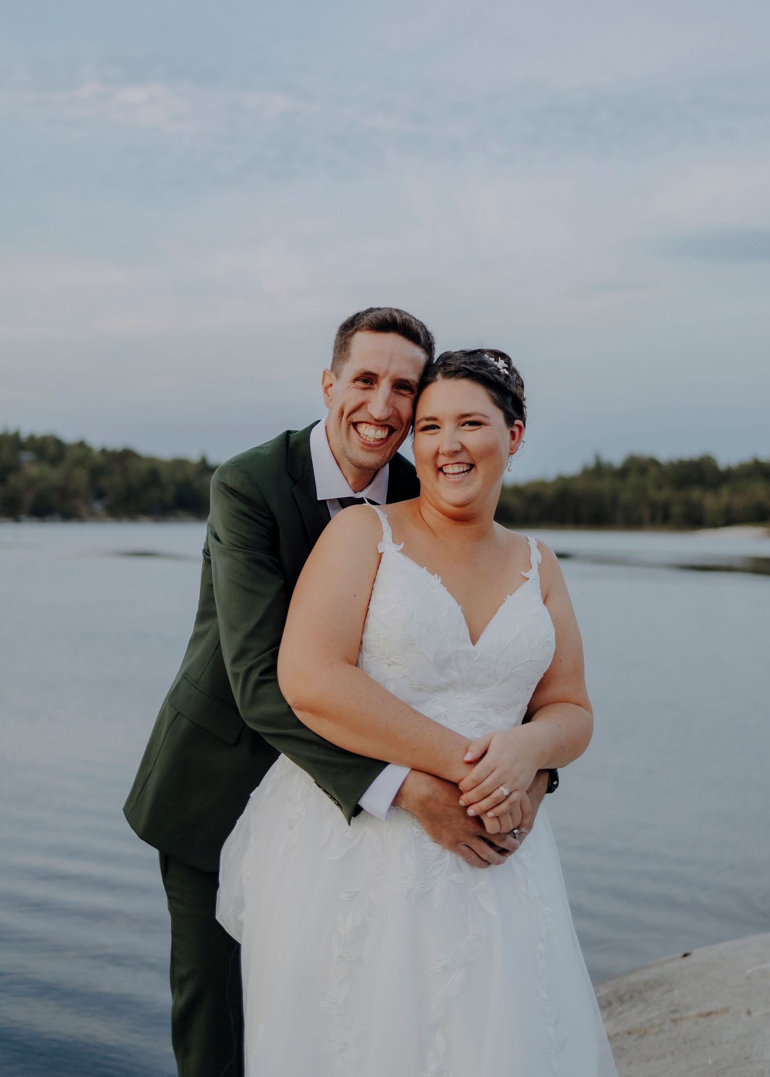 A couple shares a joyful moment by the lake during their wedding photoshoot in matching white dress and dark suit.