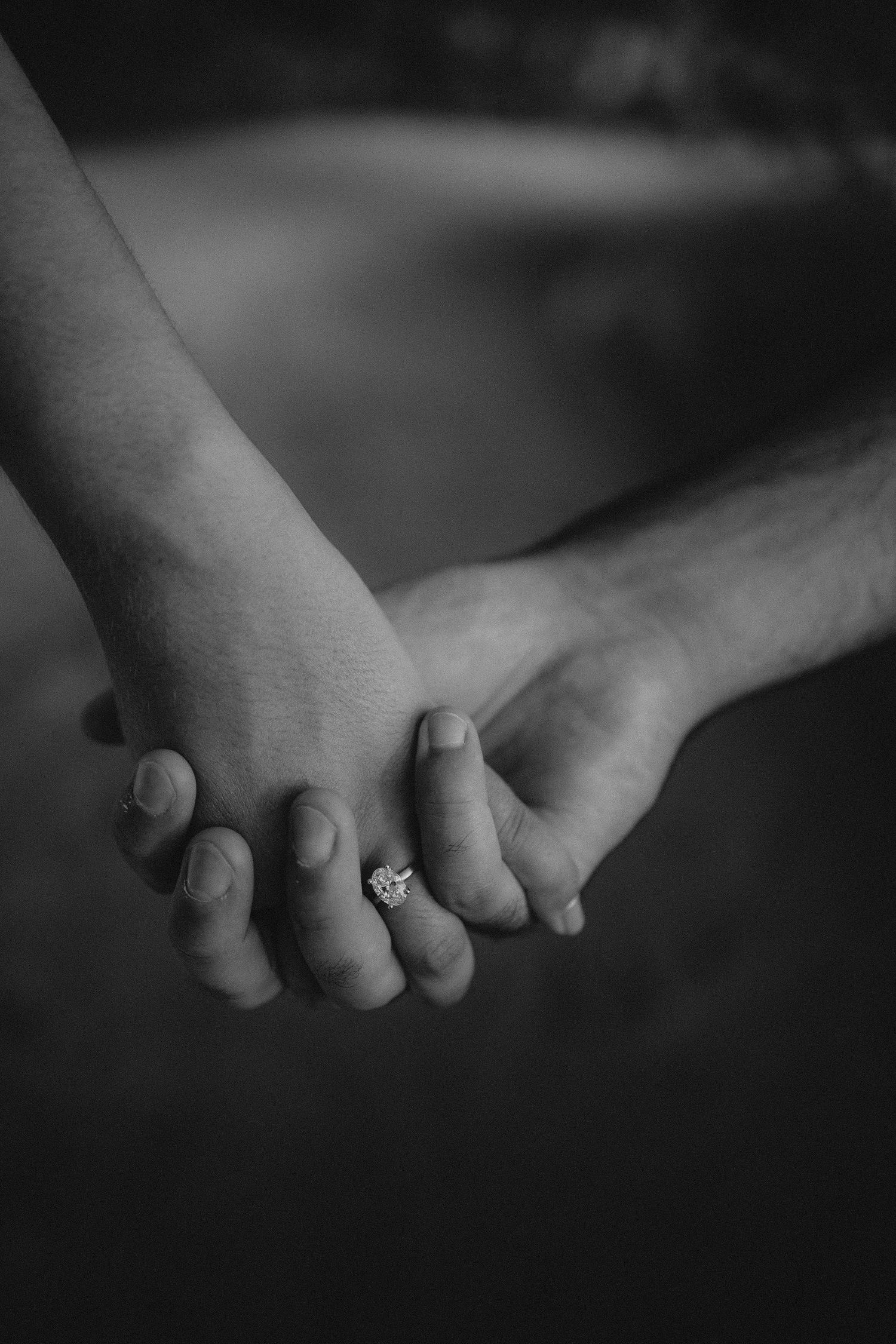 Close-up black and white photo of two hands clasping together with an engagement ring visible.
