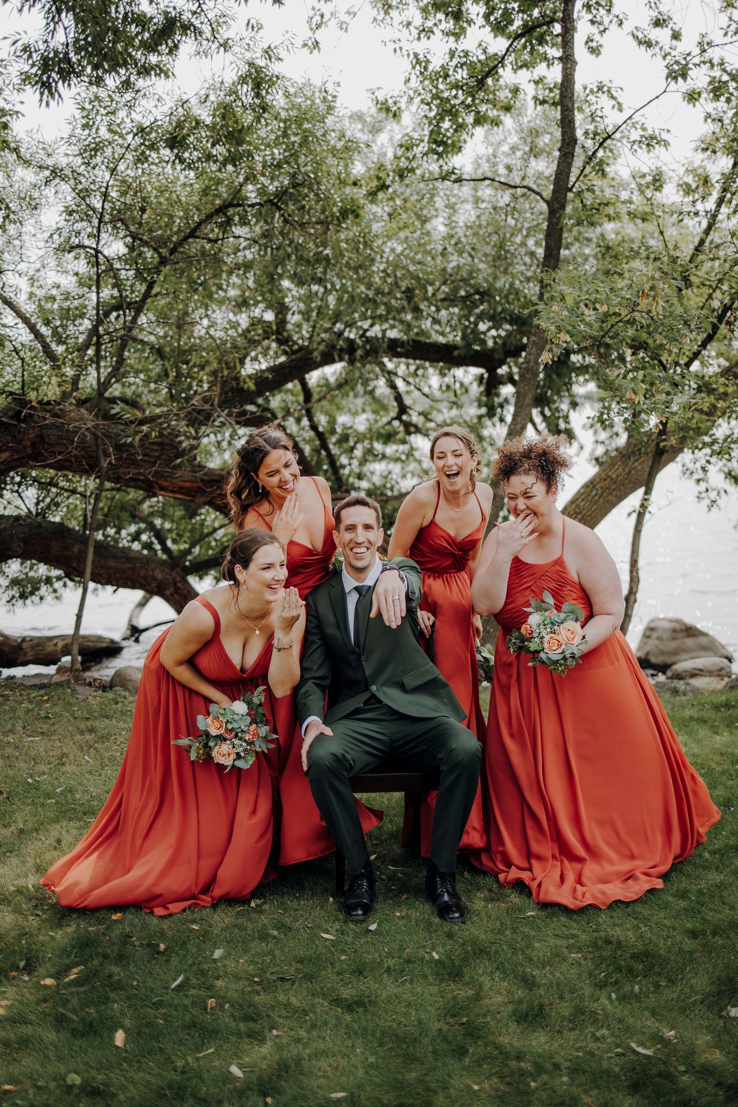 Wedding party in red dresses poses playfully with someone in a dark suit under a large tree.