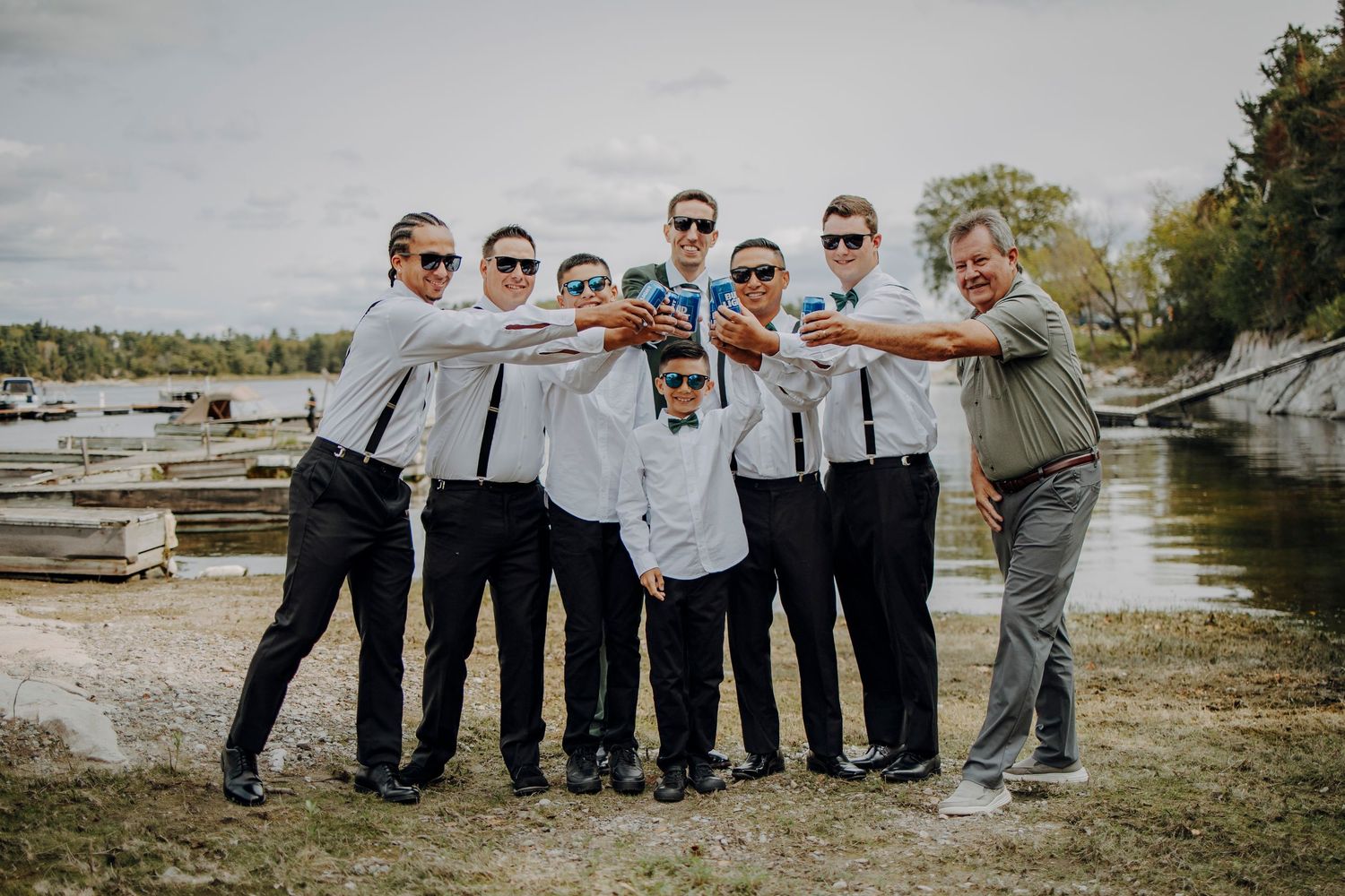 Groomsmen in matching light blue shirts and khaki pants pose playfully for wedding photos by the water.