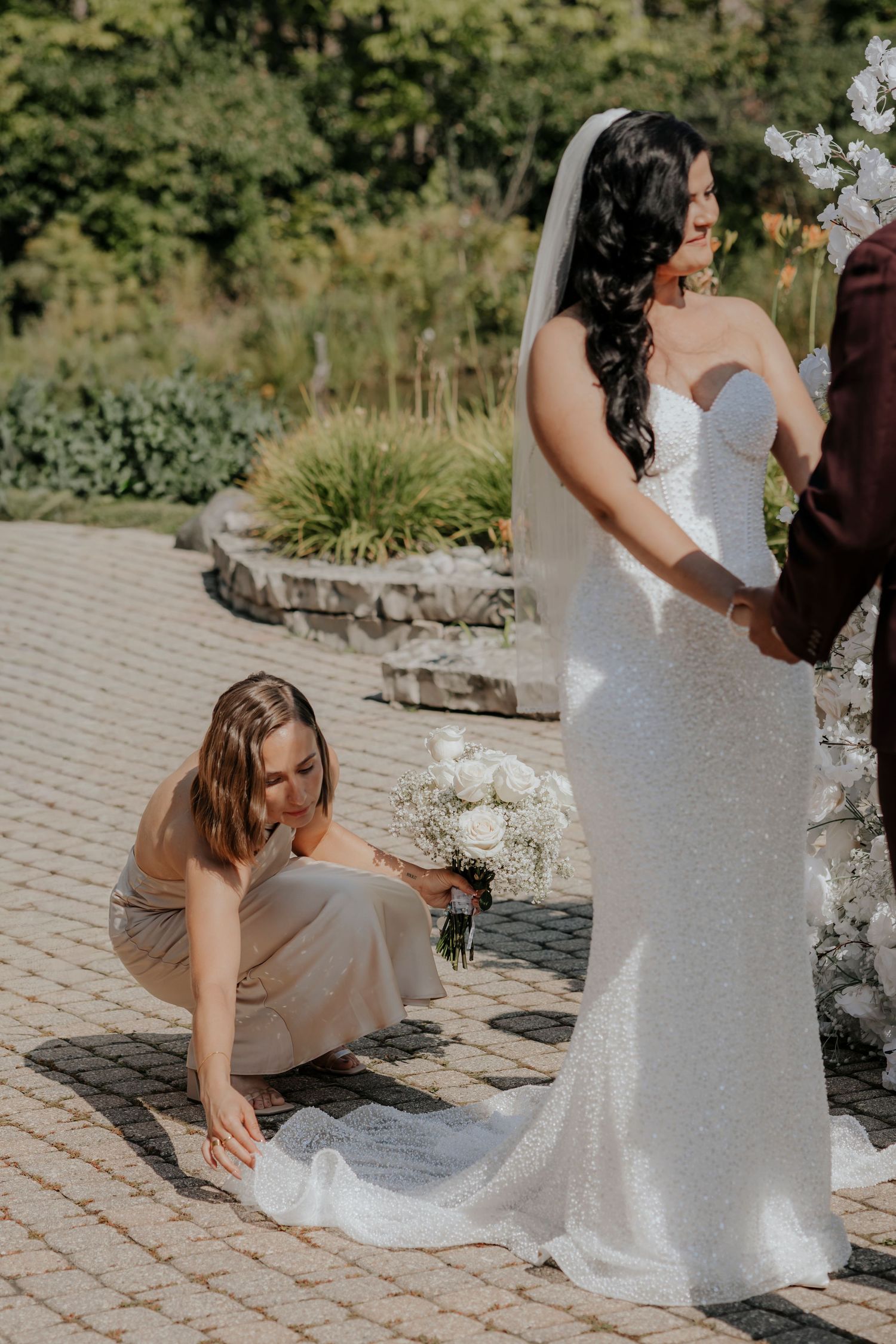 Helper adjusts the train of a bride's fitted white wedding dress on a stone patio.