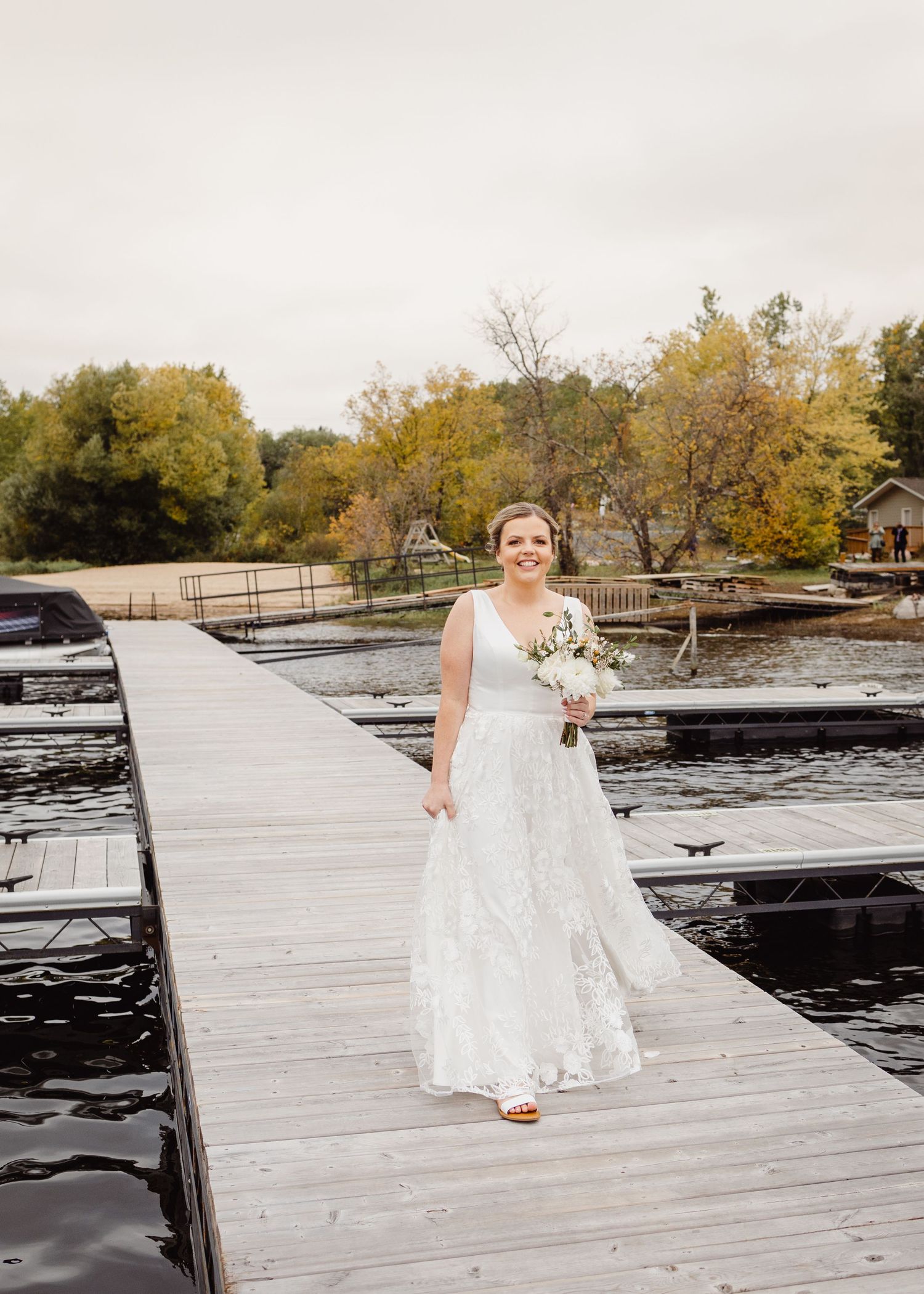 A bride in a lace wedding gown walks along a wooden dock surrounded by autumn trees and water.