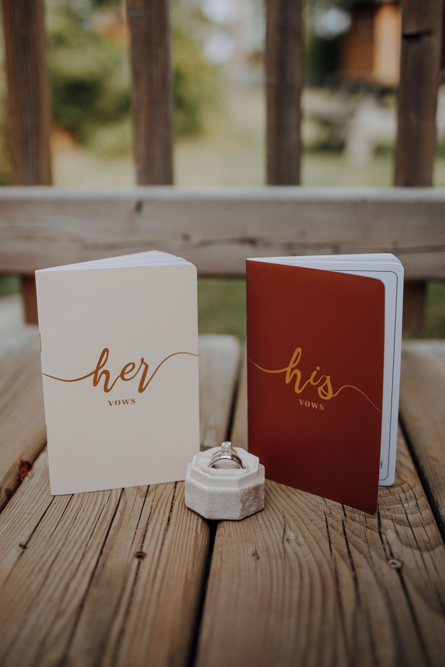 Wedding vow books and ring holders arranged on wooden deck with gold lettering.