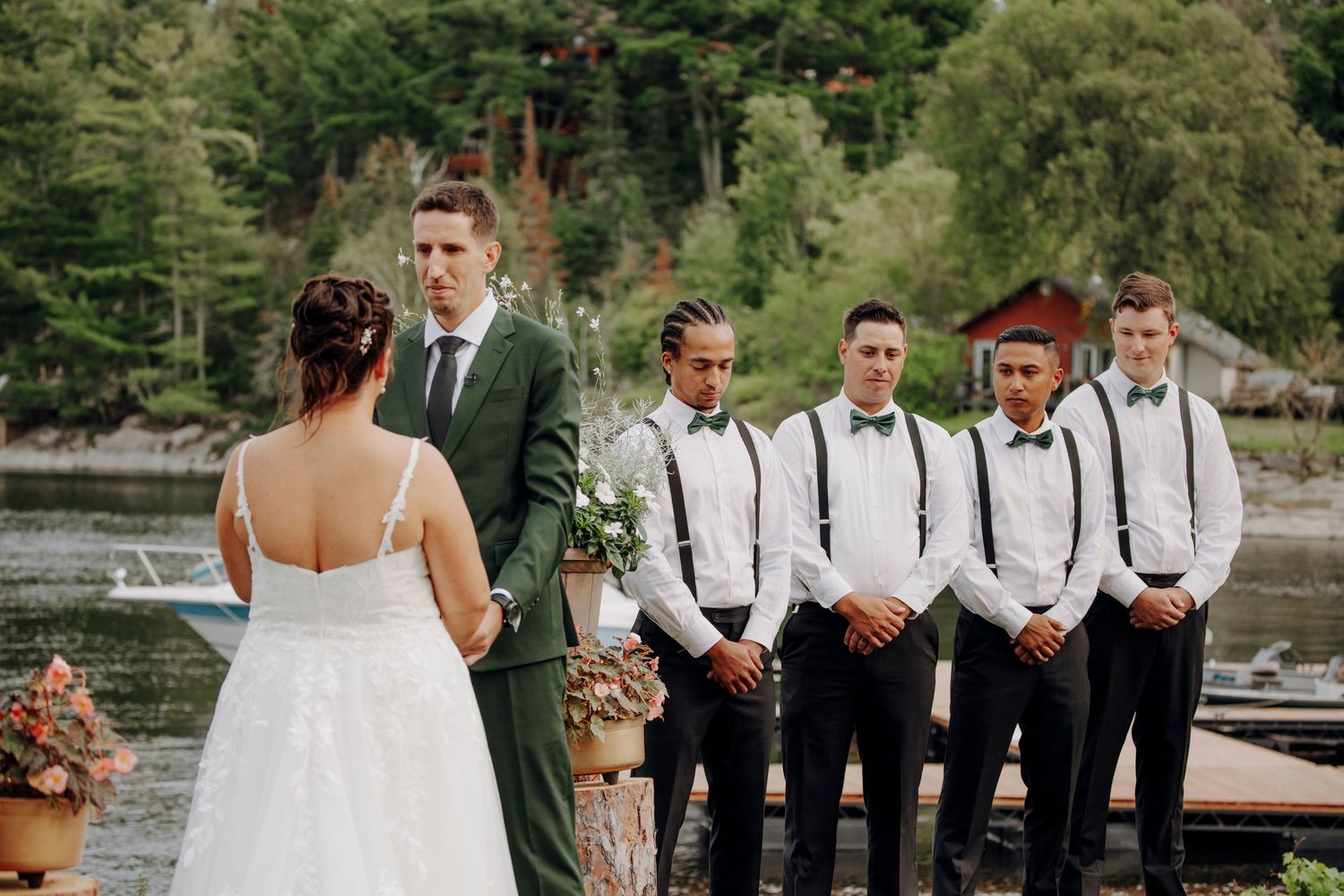 Wedding party in formal attire poses outdoors with groomsmen in suspenders and bow ties against natural backdrop.