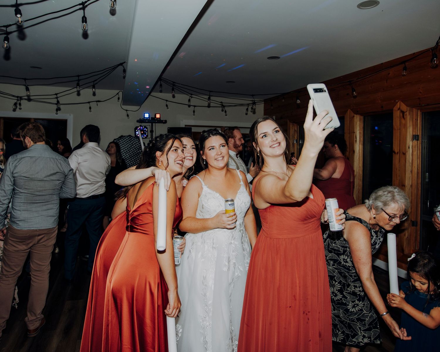 Wedding reception scene with string lights overhead and guests in coral-colored dresses mingling.