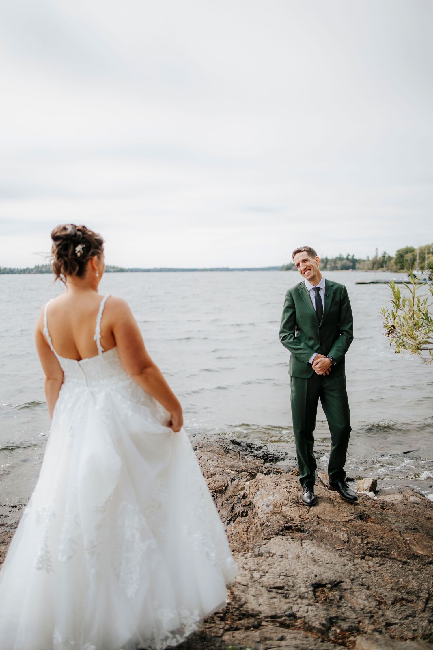 First look wedding moment between couple by waterfront with cloudy sky backdrop.