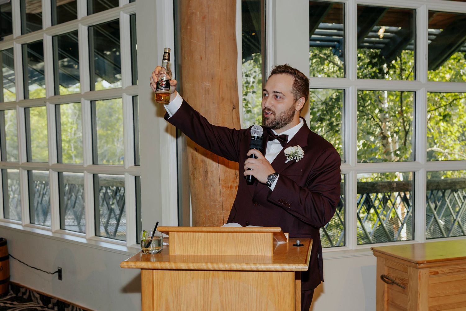 A person in a burgundy suit raises a drink in celebration inside a windowed room.