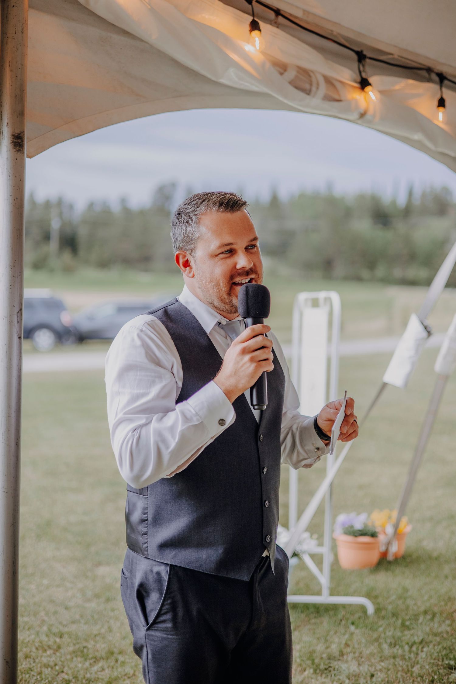 A speaker in a grey vest delivers a speech under a white tent at an outdoor event.