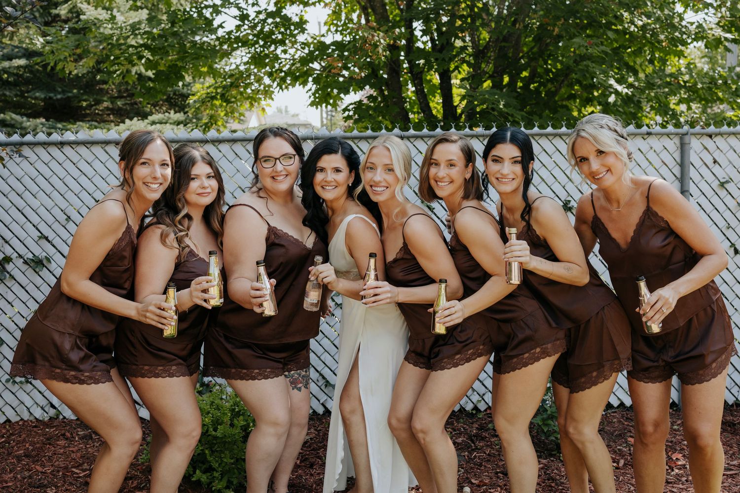 A group of bridesmaids in matching brown dresses pose together outdoors for wedding party photos.
