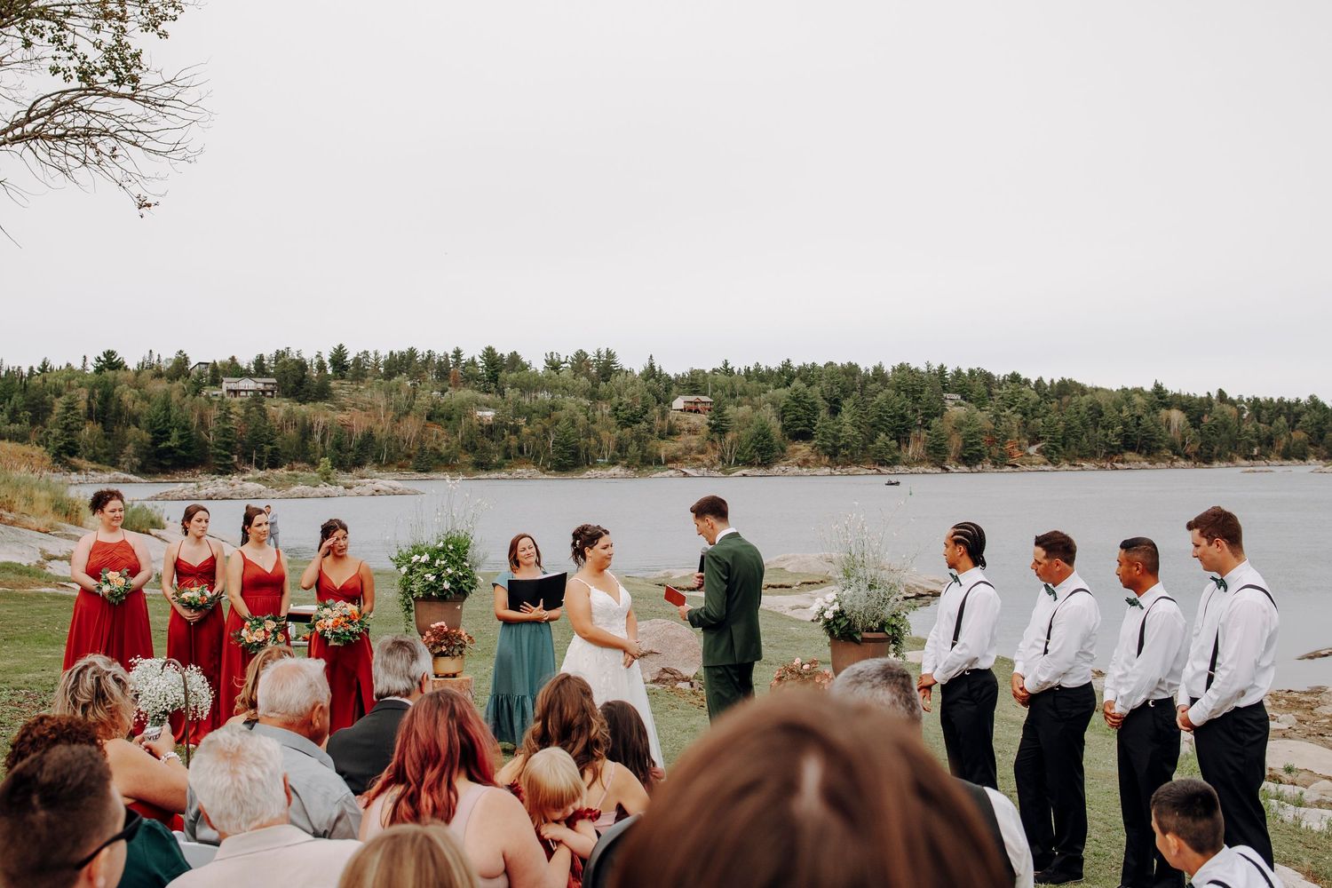 Panoramic view of an outdoor wedding ceremony setup by a lake with guests seated in white chairs.