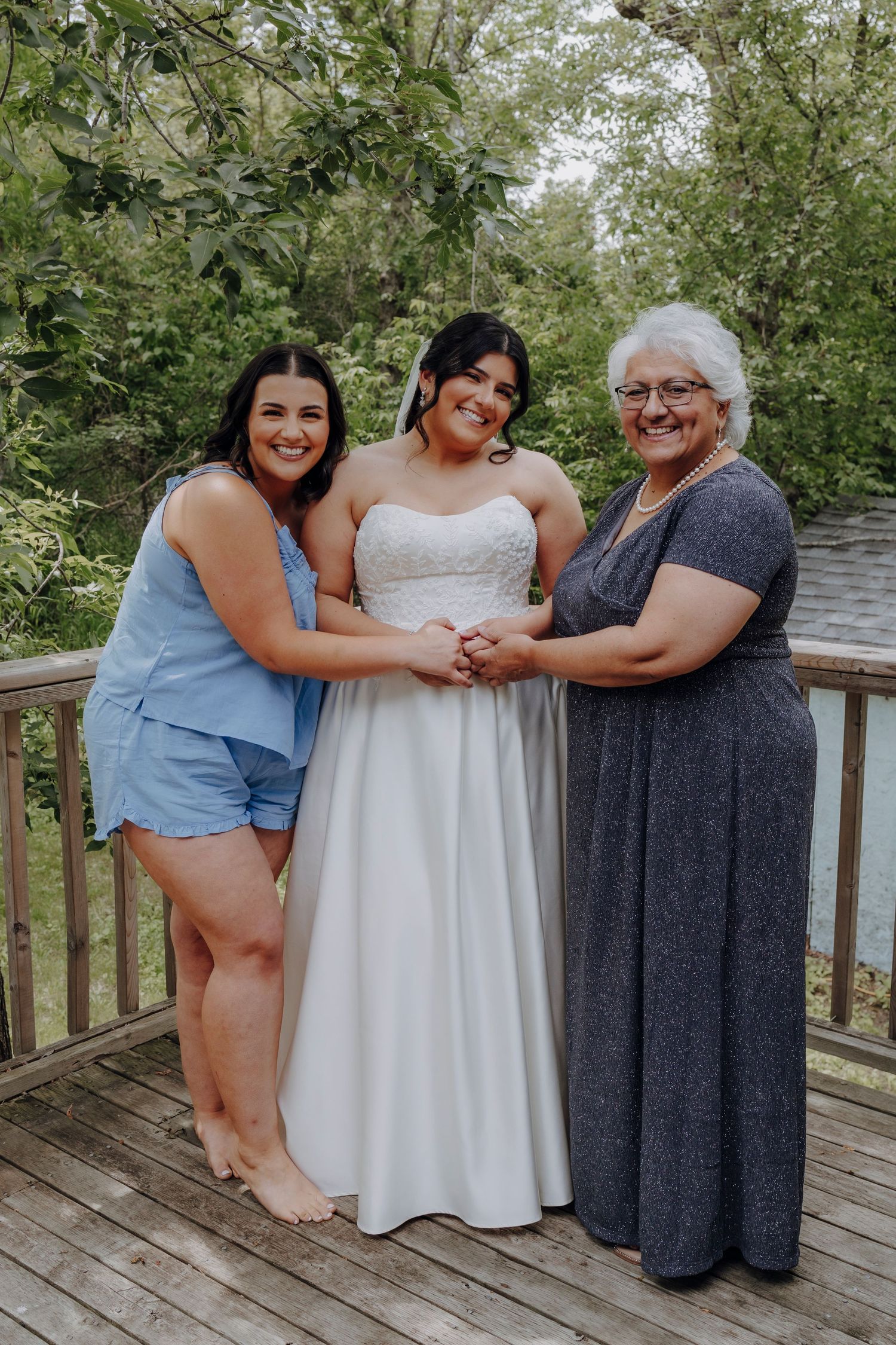 Three generations gather on a wooden deck surrounded by lush greenery wearing casual and formal attire.