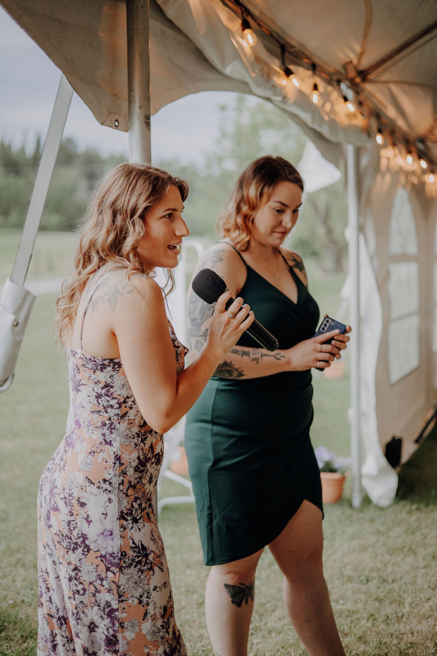 Wedding guests in floral dresses stand under a white tent with string lights during an outdoor reception.