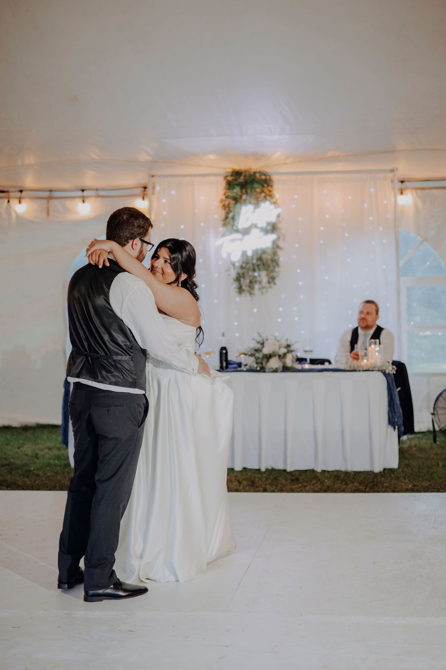 A couple shares their first dance in a white wedding tent decorated with string lights and floral arrangements.