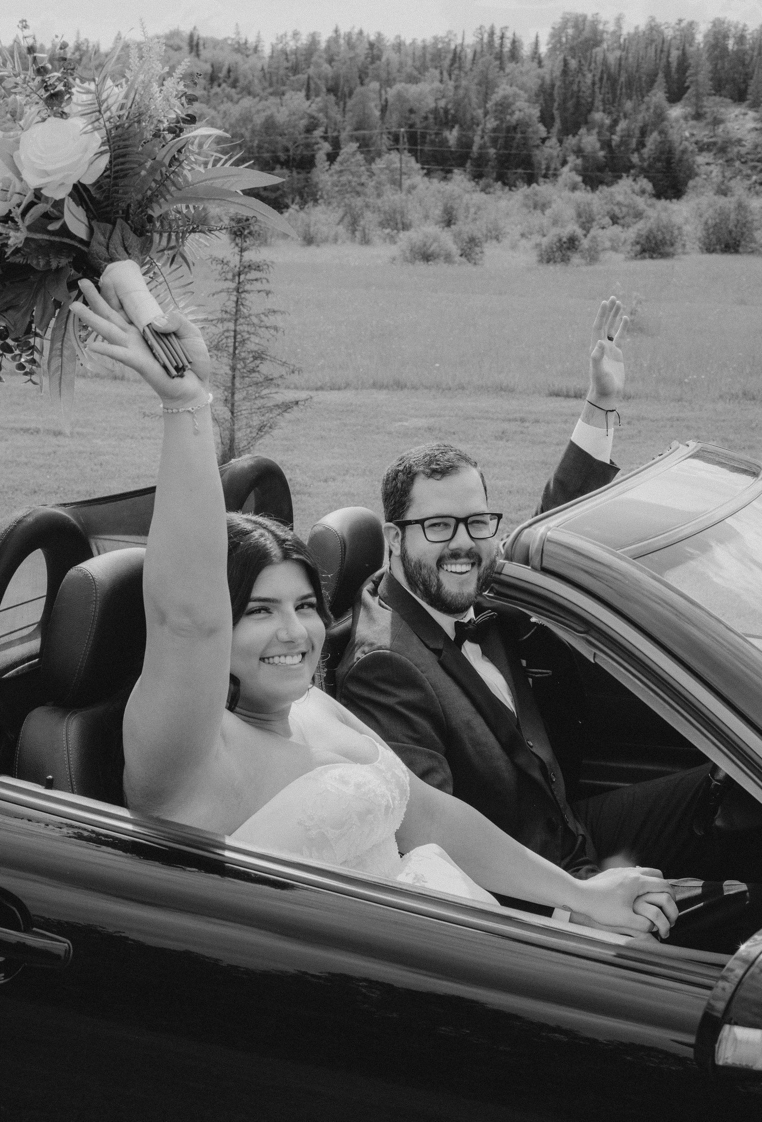 A joyful wedding couple celebrating in a convertible car with raised arms.