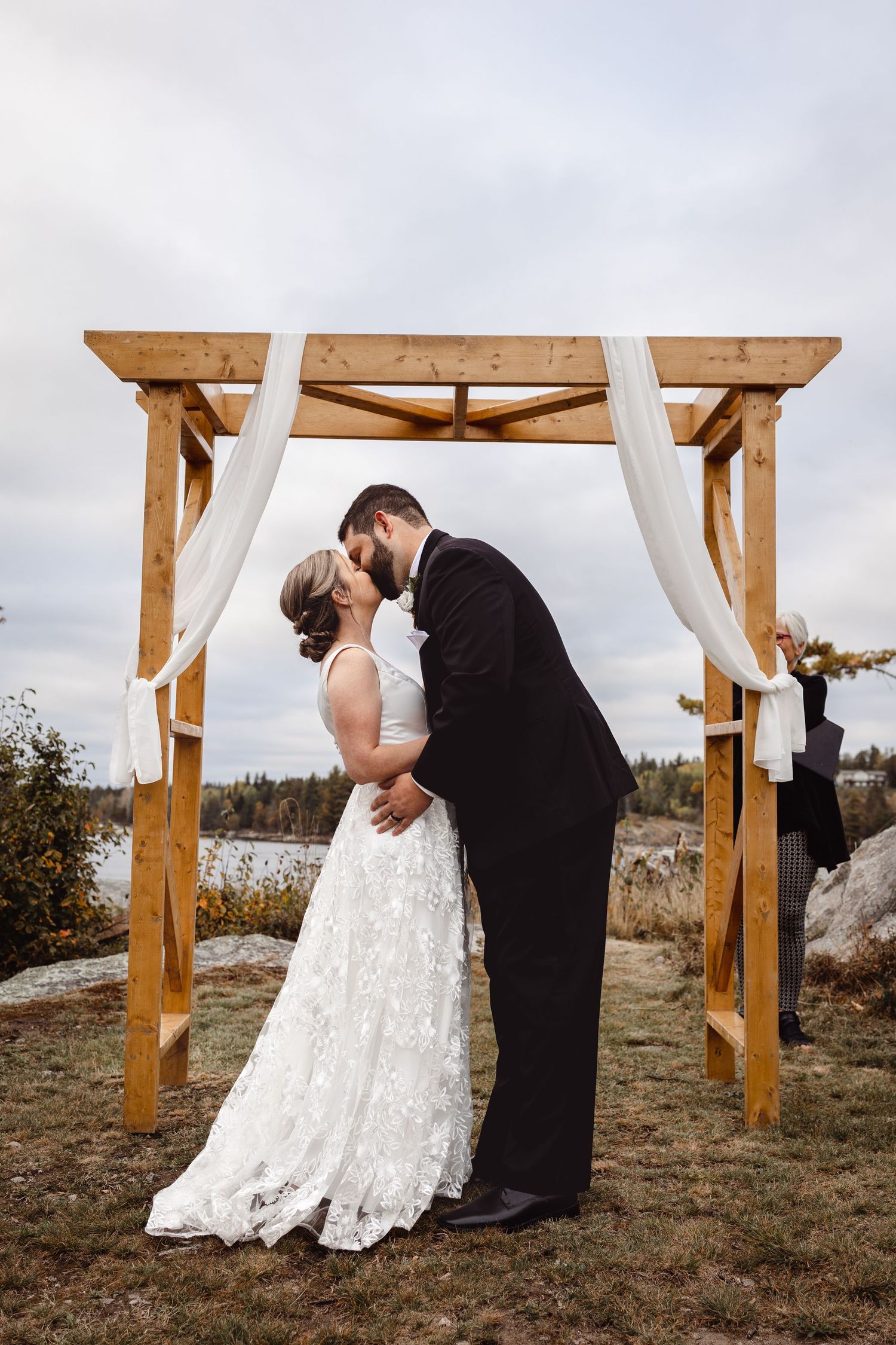 A couple shares their first kiss under a rustic wooden arch with white fabric draping during an outdoor lakeside wedding ceremony.