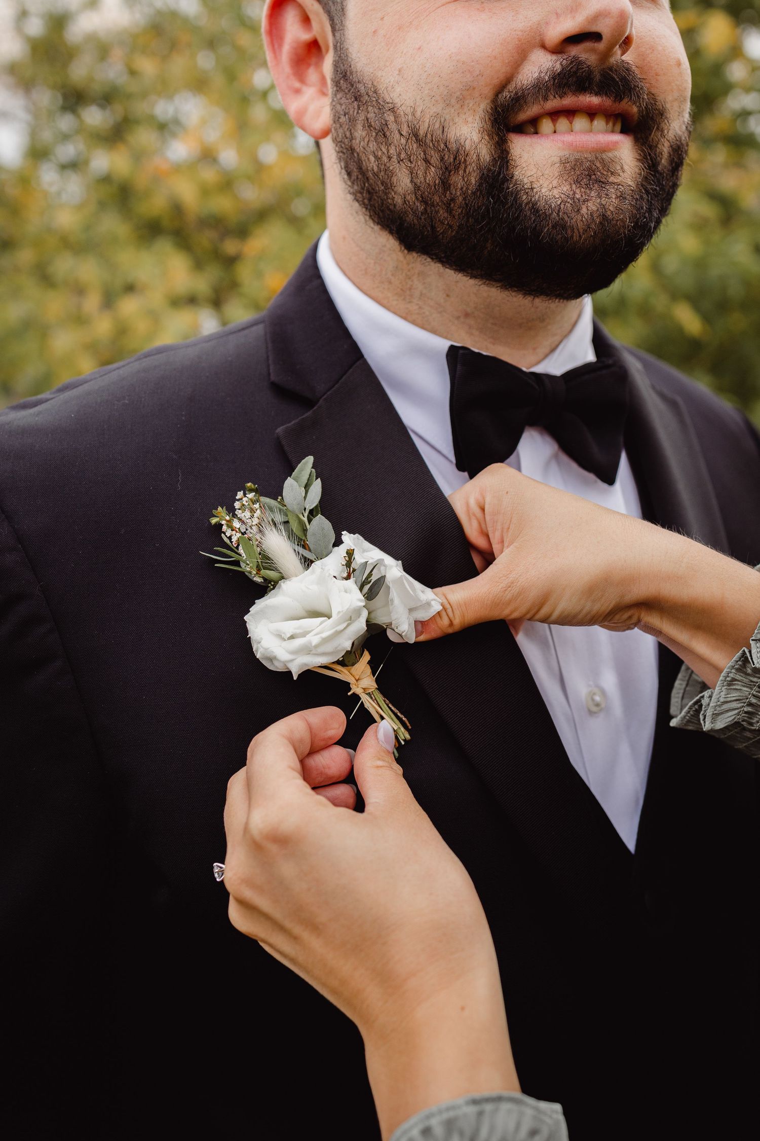 A close-up shot of hands pinning a white rose boutonniere on a black tuxedo lapel.