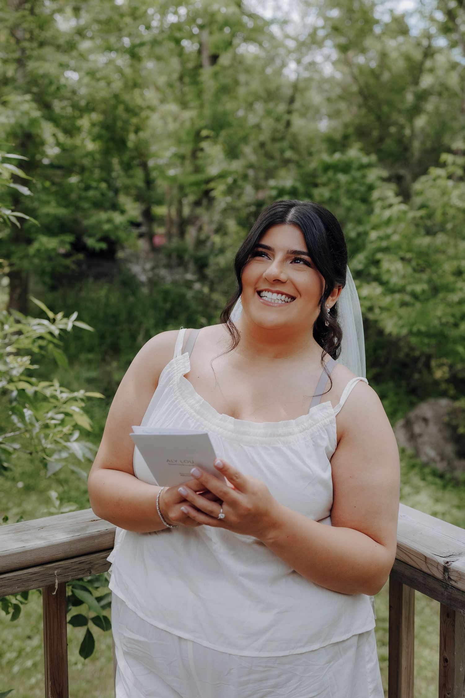 Person in a white sleeveless top sits on wooden deck surrounded by greenery while using a digital tablet.