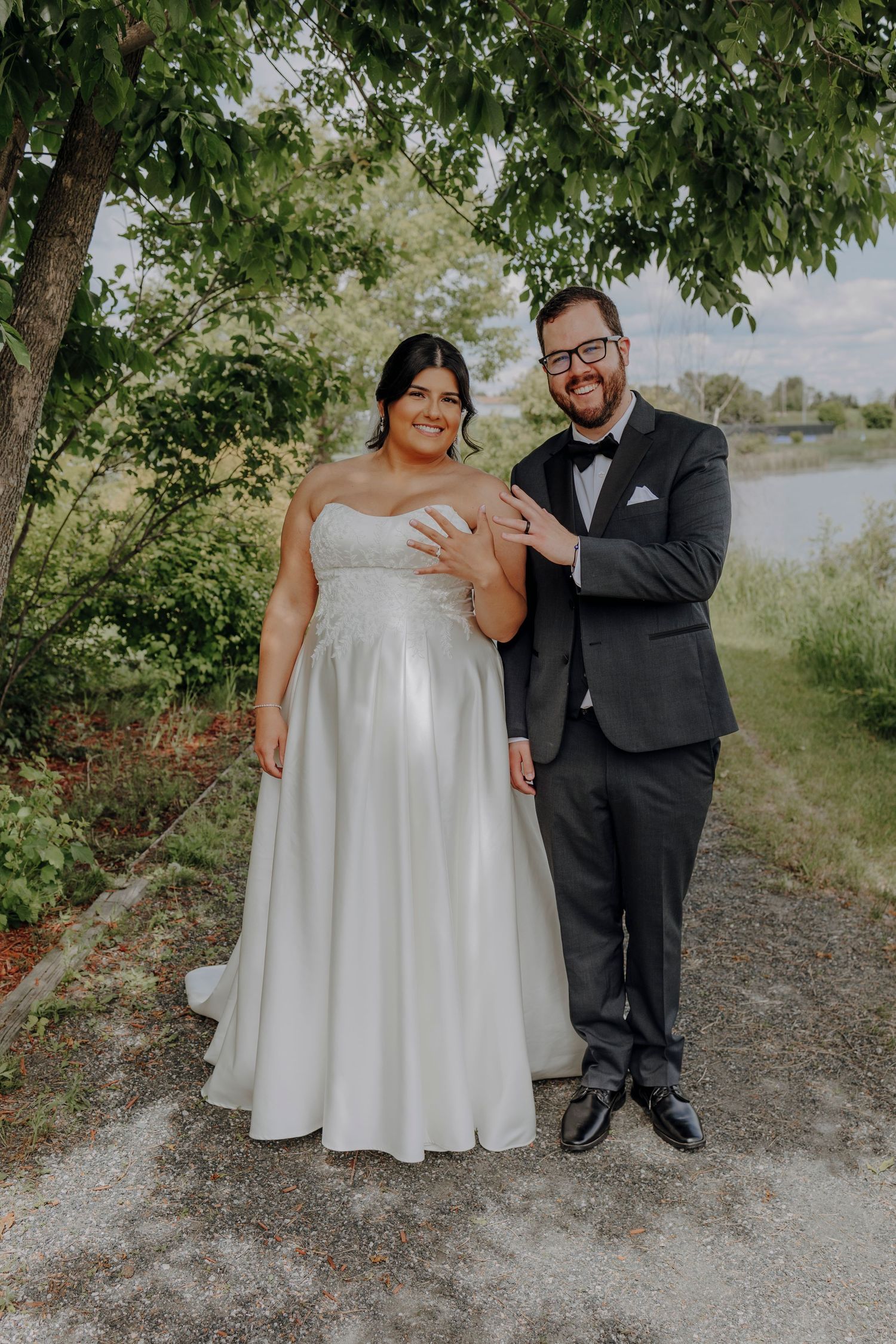 Wedding couple poses casually on gravel path with trees and water in background.