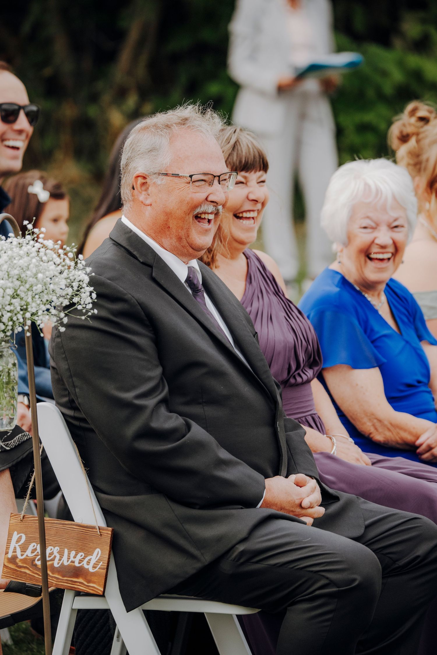 Wedding guests sharing joyful laughter while seated in wooden chairs during an outdoor ceremony.