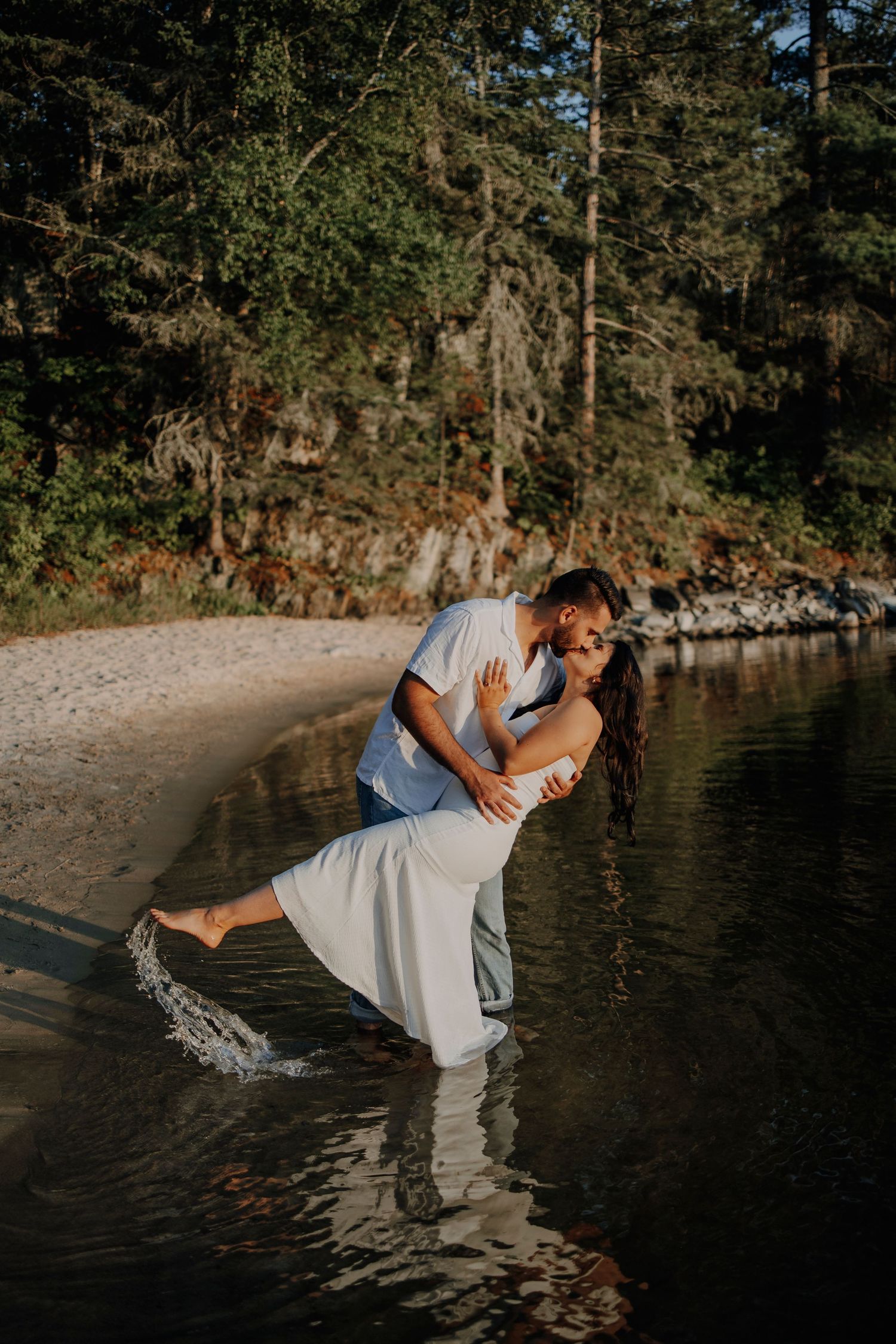 A playful romantic dip pose by the water's edge with pine trees in the background.
