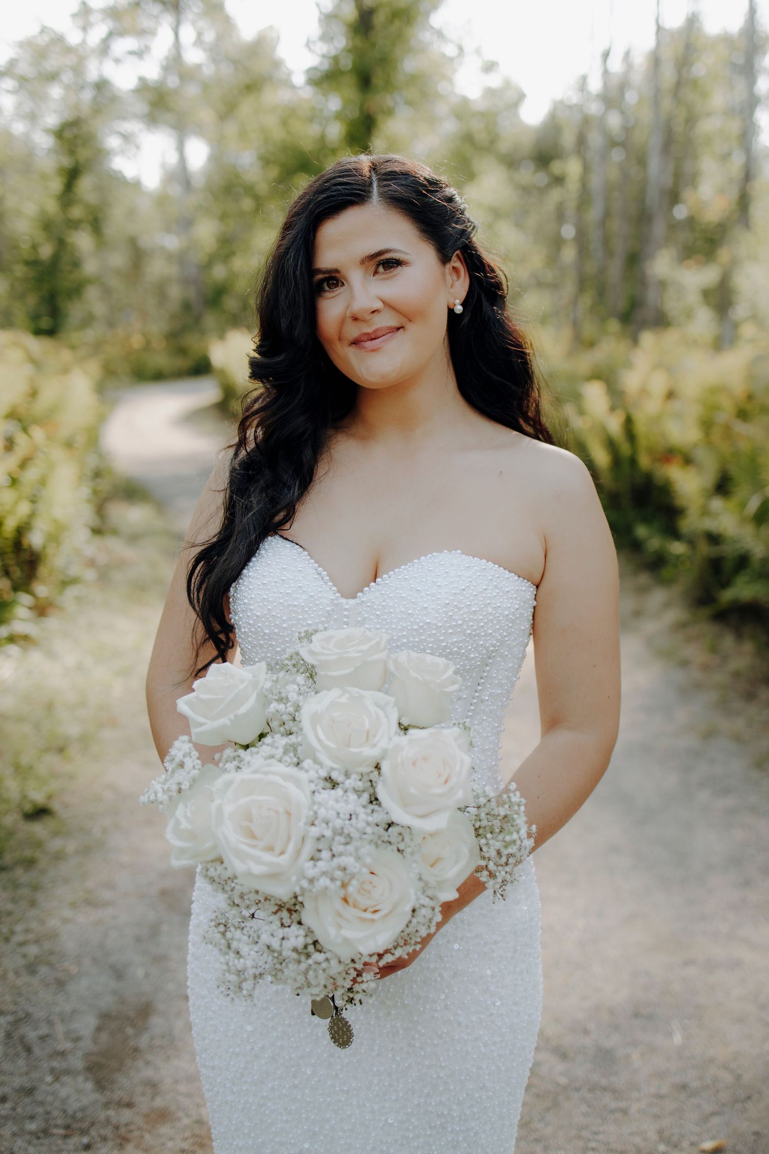 A bride in a white strapless dress holds an elegant bouquet of white roses and baby's breath against a natural outdoor setting.