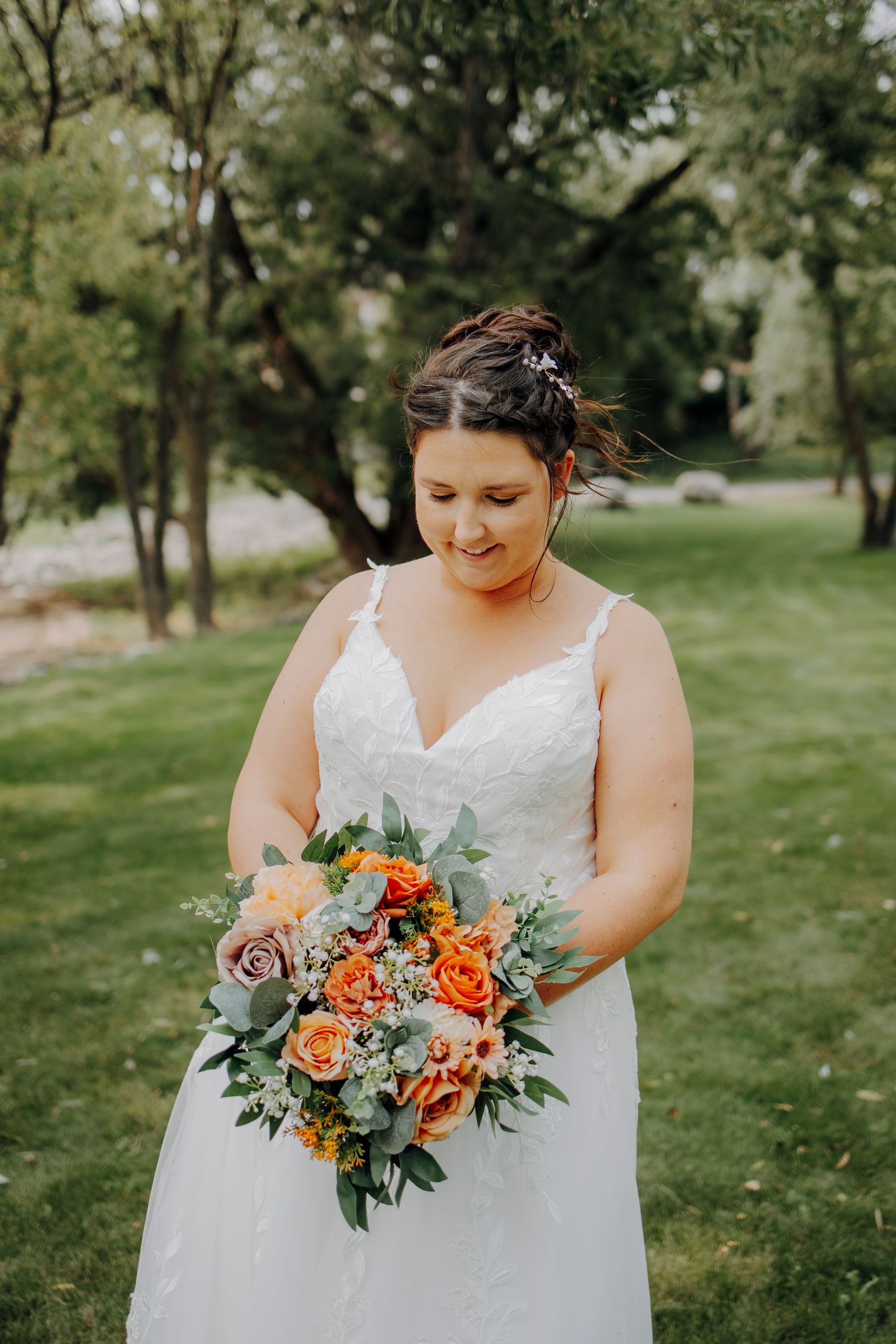 A bride in a white lace dress holds a vibrant coral and peach rose bouquet in a garden setting.