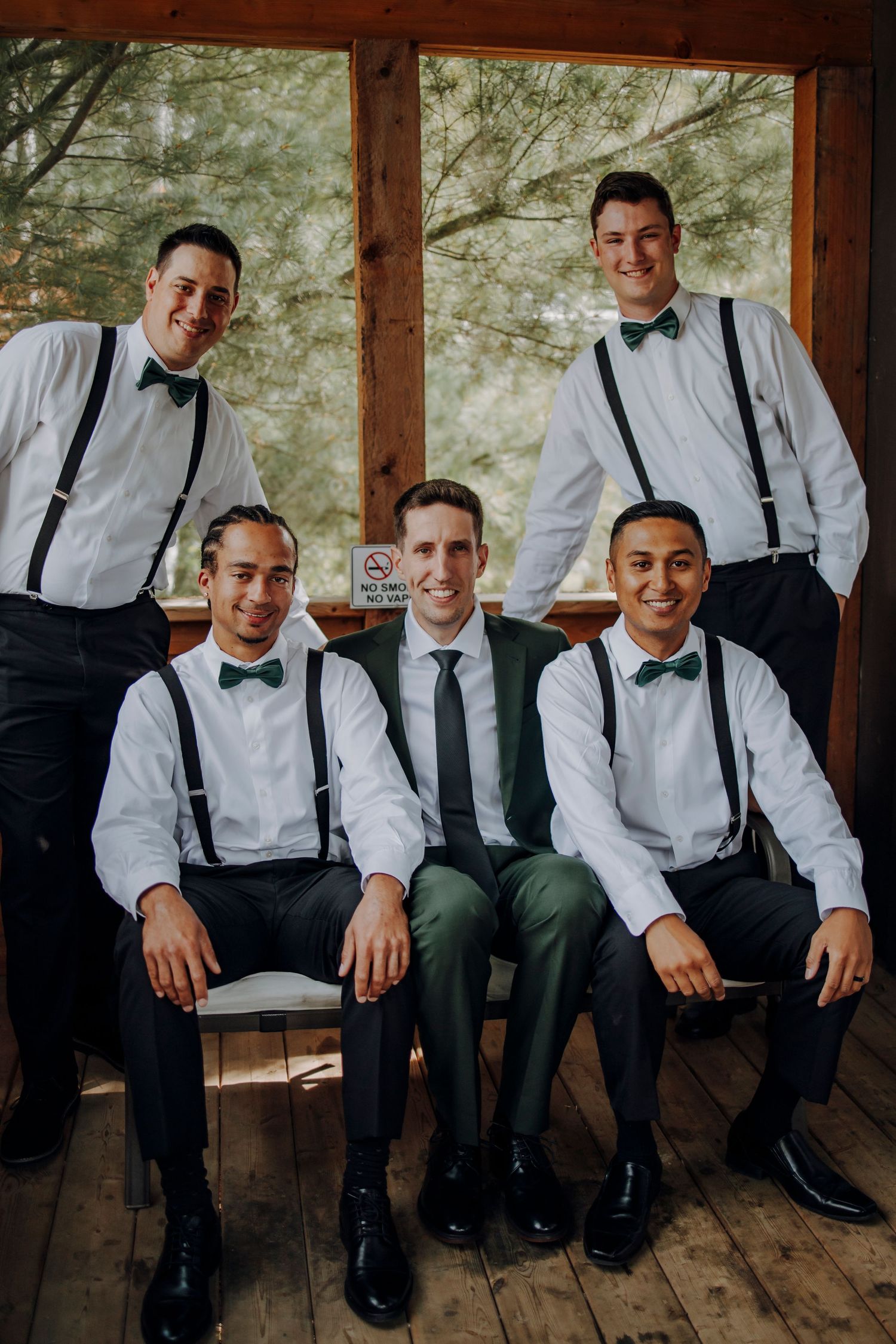 Groomsmen in white shirts and suspenders sitting together on wooden bench for wedding photos.