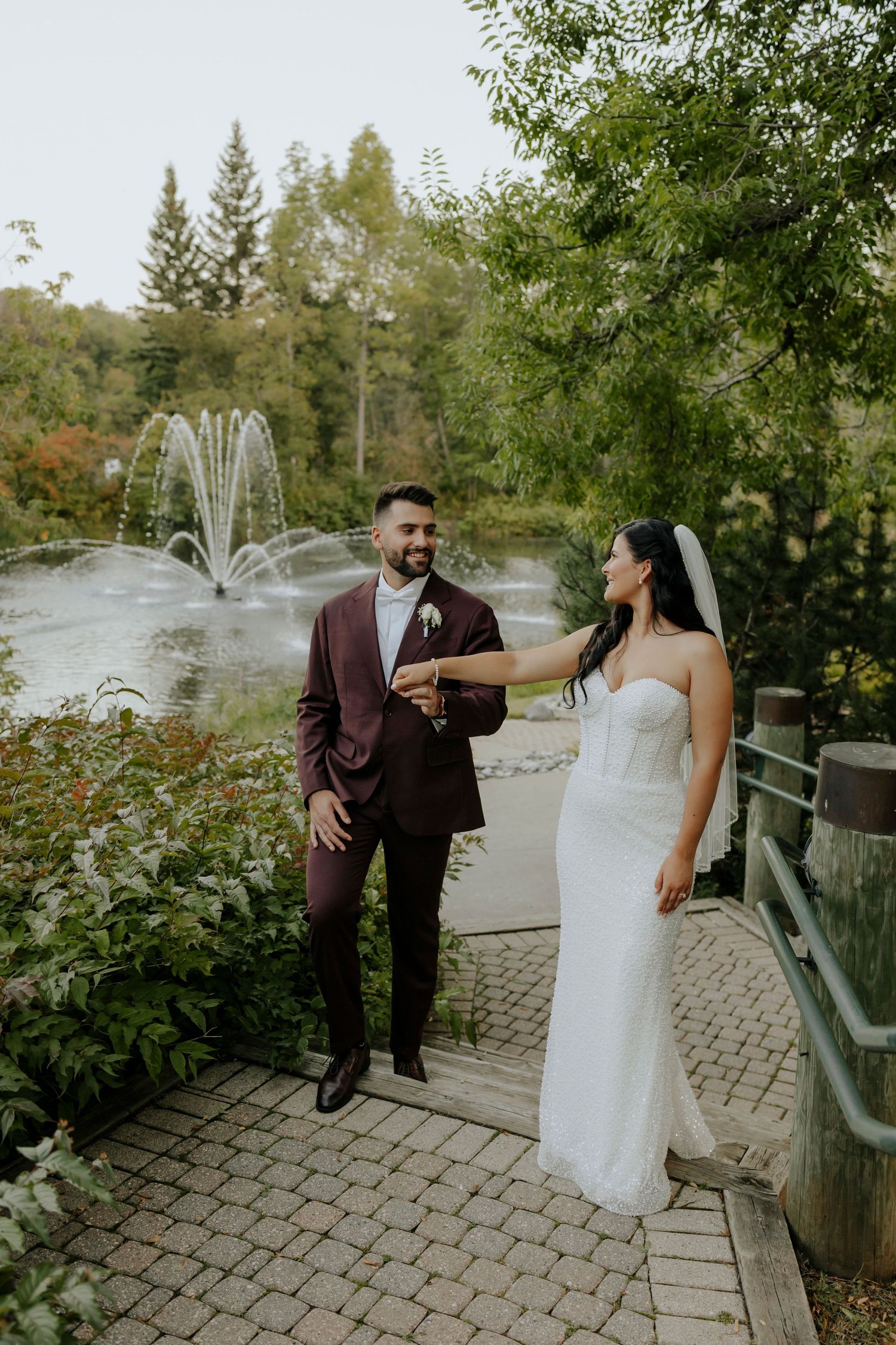 A couple shares a joyful moment on a stone path with a fountain and lush greenery in the background.