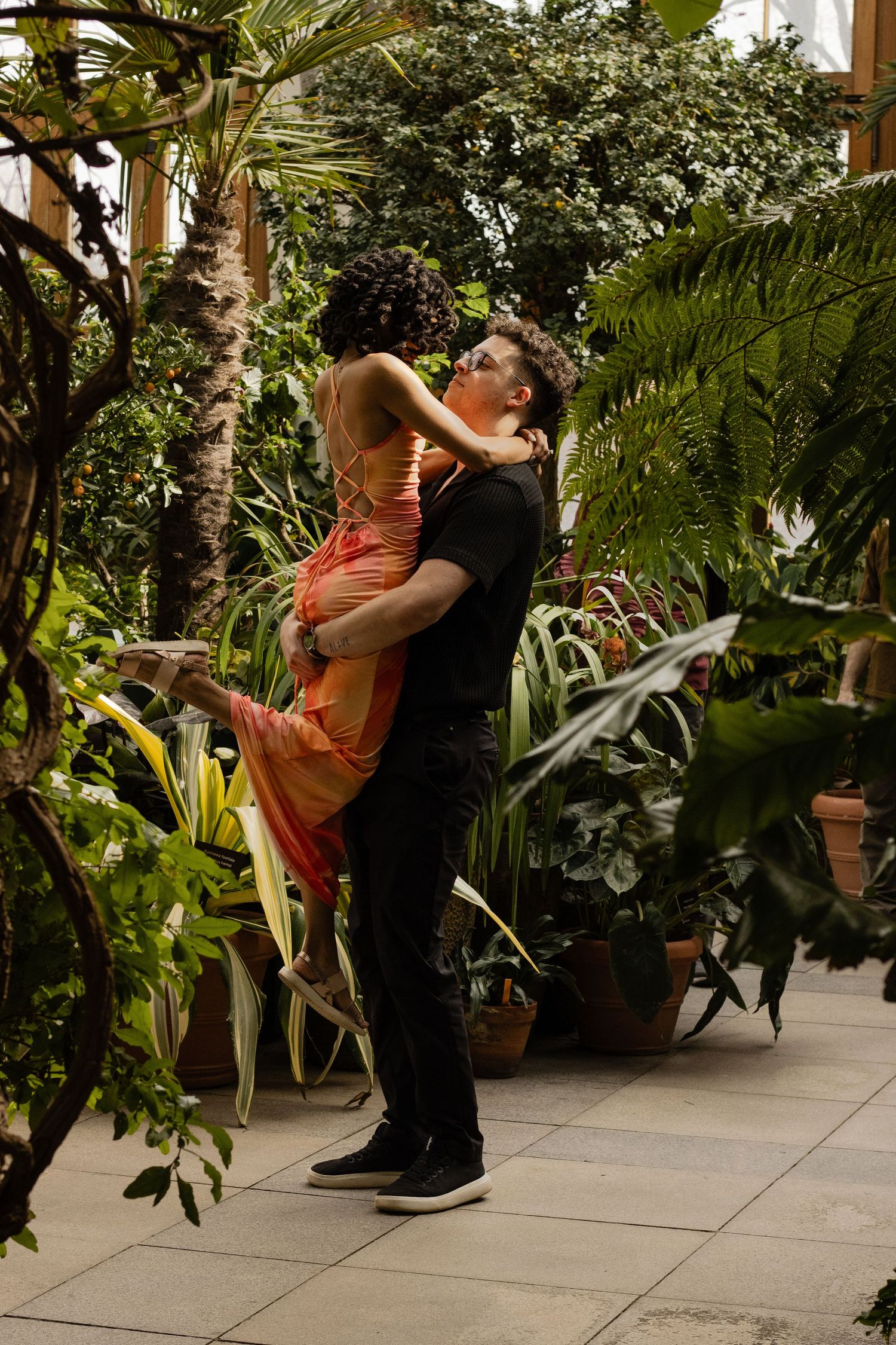 Man lifting woman in an orange dress in a lush indoor botanical garden setting.