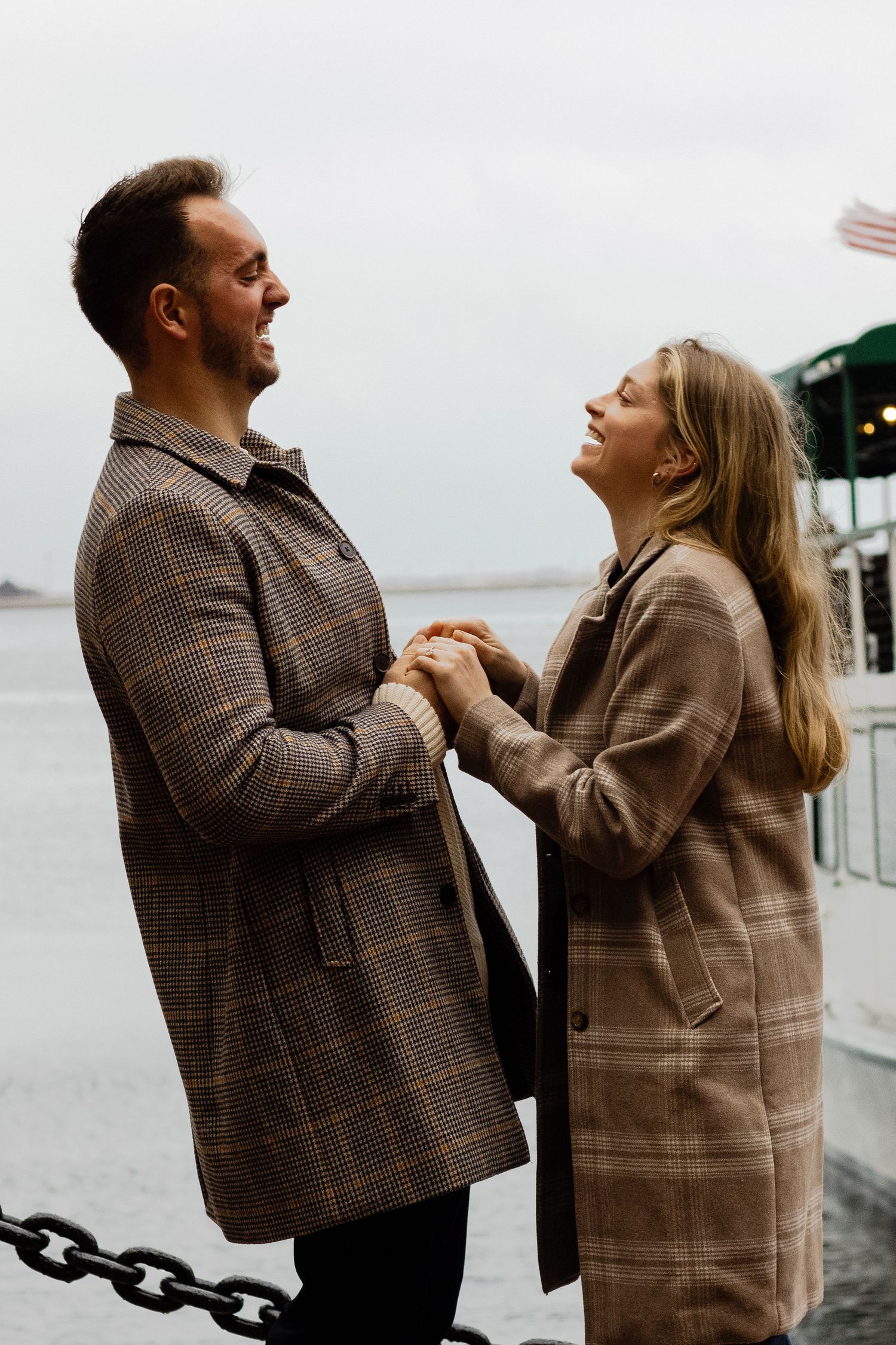 A joyful couple holds hands and gazes at each other near a waterfront on a cloudy day.
