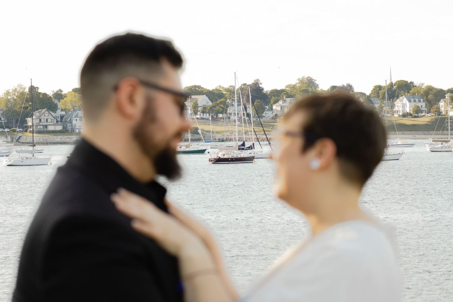 Couple sharing a romantic moment by a scenic harbor with sailboats and coastal town in the background.